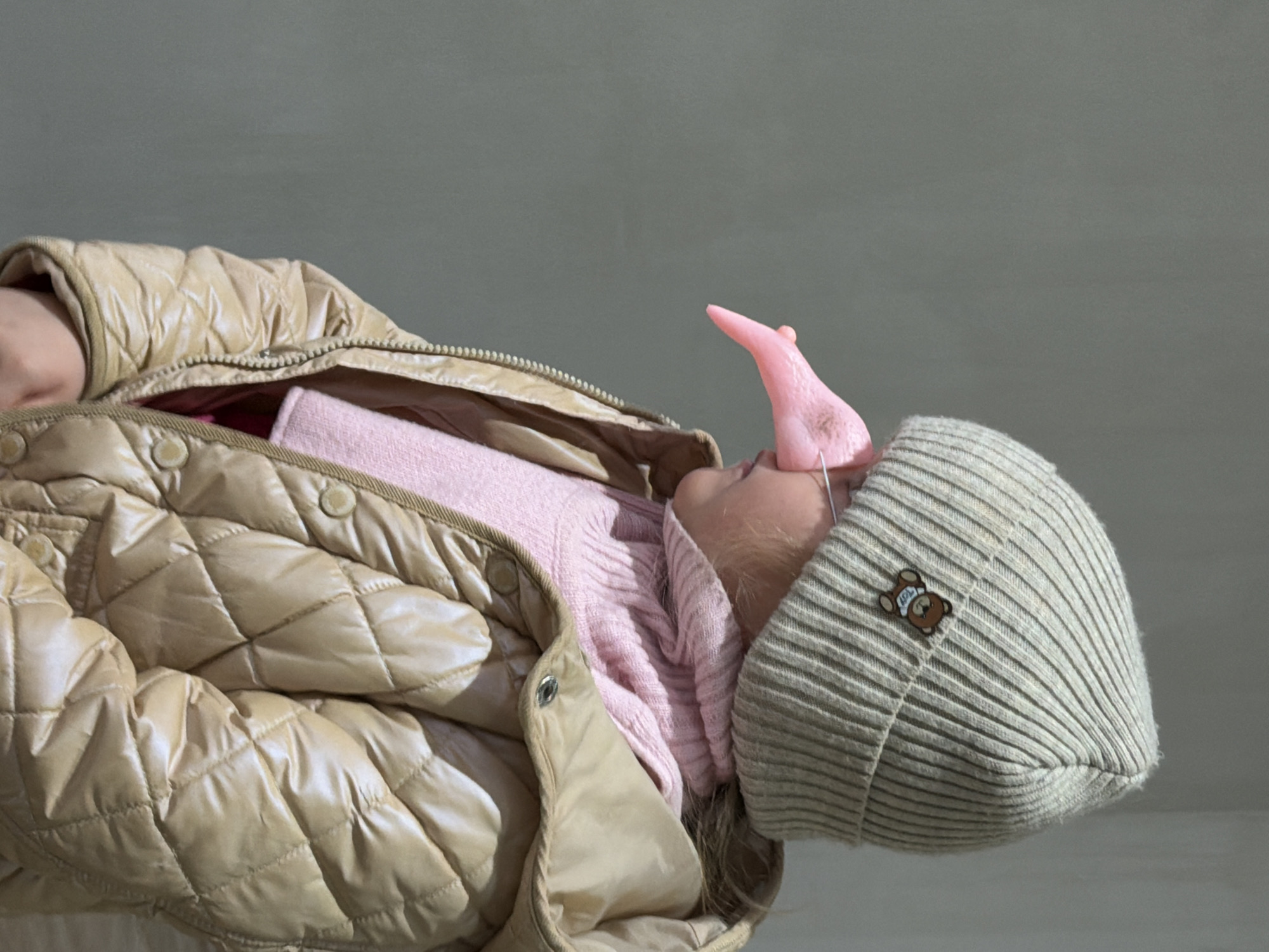 The image shows a child wearing a beige quilted jacket over a pink sweater and a ribbed knit hat with a bear applique. A pink, horn-shaped object is attached to the child's forehead. The background is a plain gray wall. The location, Chaikovsky, Russia, is not discernible from the image.