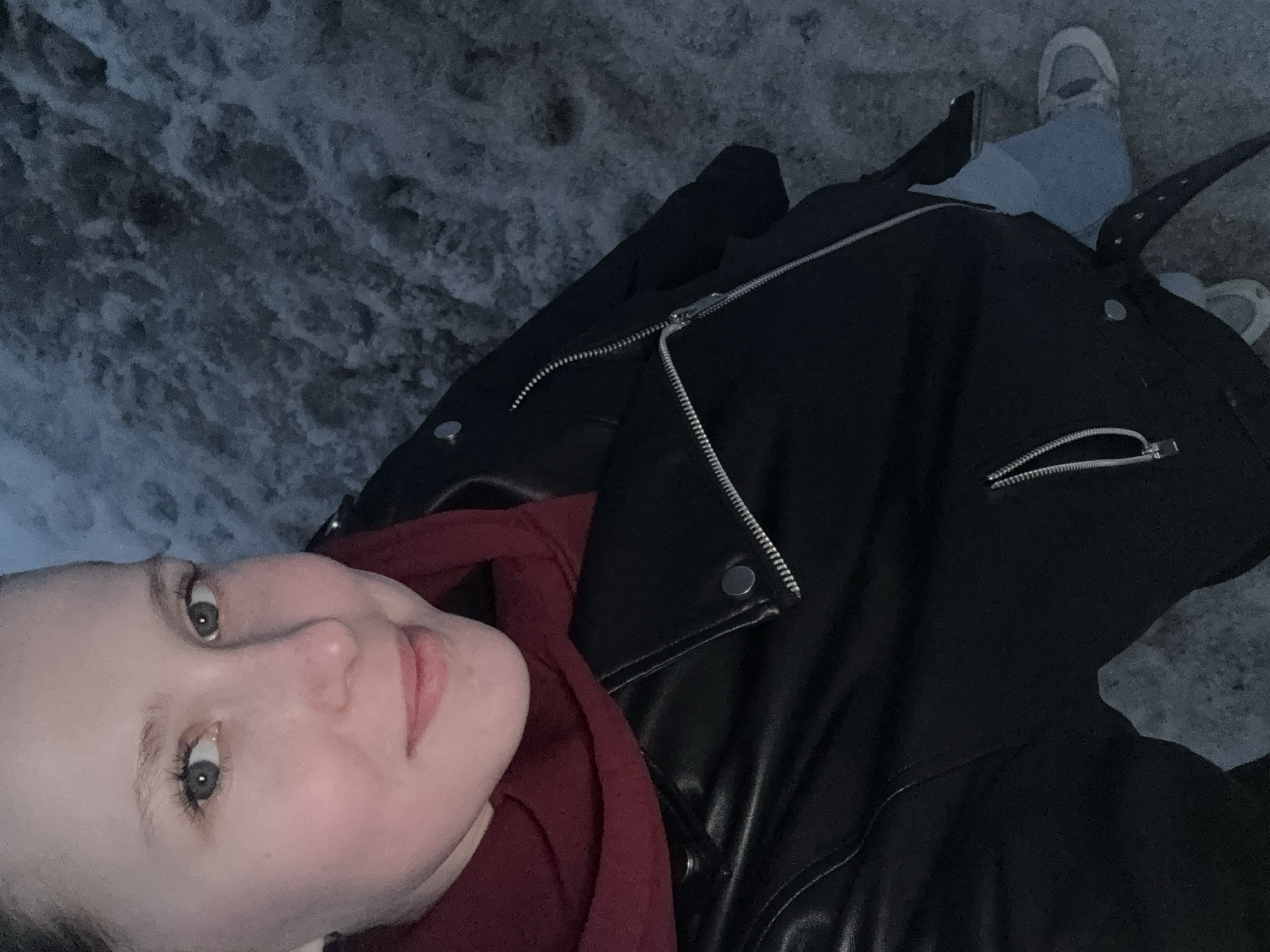 A young woman in a black leather jacket and red scarf takes a selfie. She is standing outside, presumably in the city of Chaikovsky, Russia, on what appears to be a snowy or icy surface. The woman is wearing jeans and white sneakers.