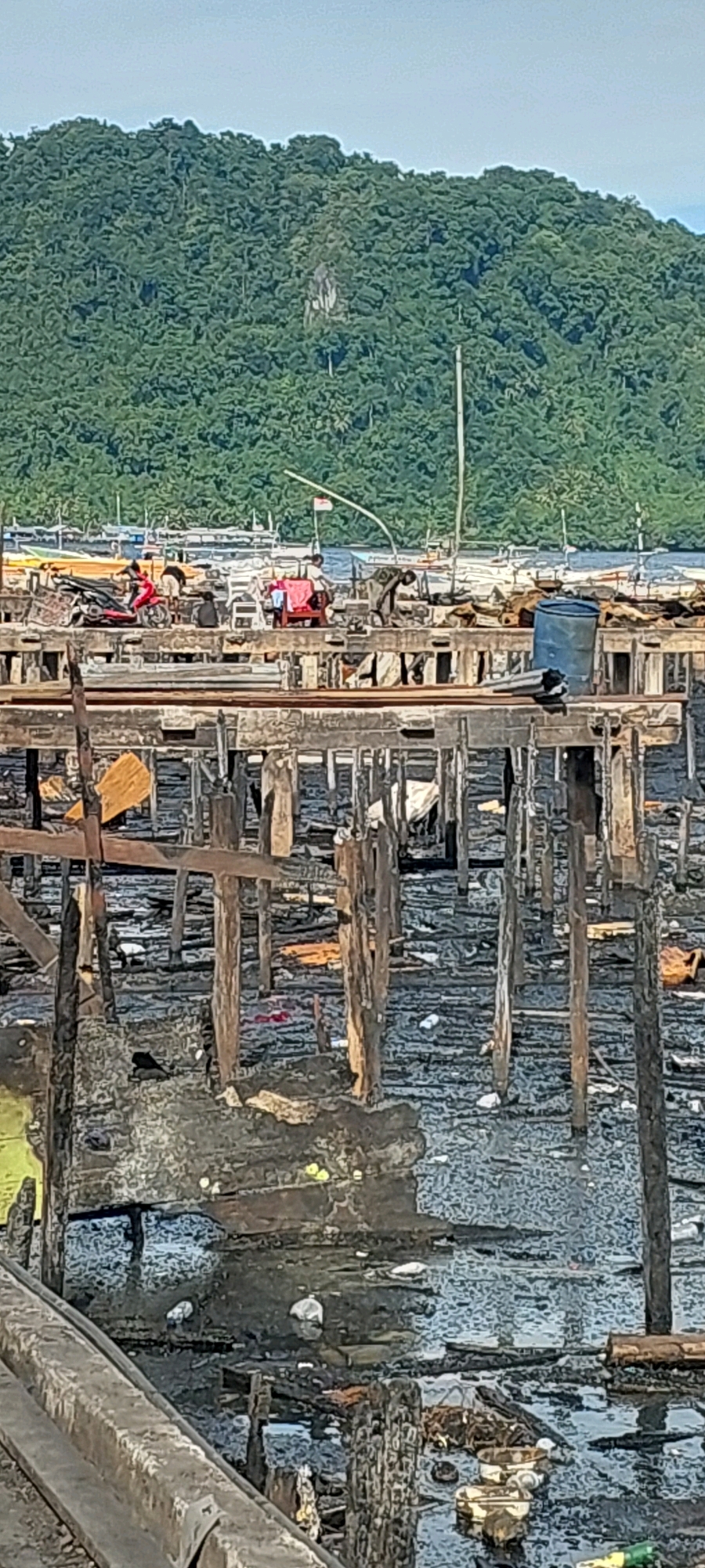 The image depicts a scene of destruction and disarray in what appears to be a waterfront community, potentially within Jayapura, Indonesia. In the foreground, wooden structures, likely part of stilt houses or a pier, are in a state of disrepair, with many posts broken and the ground below covered in debris and murky water. Scattered items, including what could be remnants of household goods and refuse, are visible amidst the water and damaged structures.
Further back, a raised wooden platform is visible, supporting various objects. A red motorcycle is parked on this platform, and several people are present, one appearing to be on the motorcycle. Another individual is seen near some red chairs and clothes that are hanging, possibly to dry. A blue plastic drum is also present on this platform, along with other assorted items.
Beyond this platform, a body of water is visible, dotted with numerous boats of varying sizes. In the background, a lush, green, tree-covered hillside rises prominently, suggesting a natural landscape adjacent to the urban or semi-urban settlement. The sky above is a light blue, with a few faint clouds. The overall impression is of a community impacted by significant damage or deterioration, with signs of recent activity amidst the wreckage.