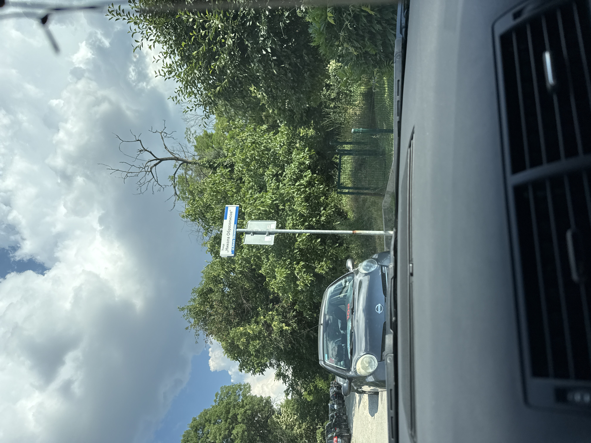 The image shows a view from inside a vehicle, looking out through the windshield. The scene is outdoors, with lush green trees and a cloudy sky. A street sign is visible, indicating a street name in Cyrillic script: "Улица Никола Образоцов". The sign is attached to a pole and appears to be a standard street sign.
In the foreground, part of the vehicle's interior is visible, including the dashboard and an air vent. Outside the vehicle, several cars are parked in a line. One car, a dark-colored hatchback, is particularly visible in the mid-ground. The overall impression is of a street scene on a partly cloudy day, possibly in a suburban or urban area with a lot of greenery. The lighting suggests it is daytime, but the heavy clouds might indicate approaching rain or overcast conditions. There are no people clearly visible, but the presence of parked cars and a street sign suggests a populated area. The image is oriented vertically, with the sky at the top and the road at the bottom.