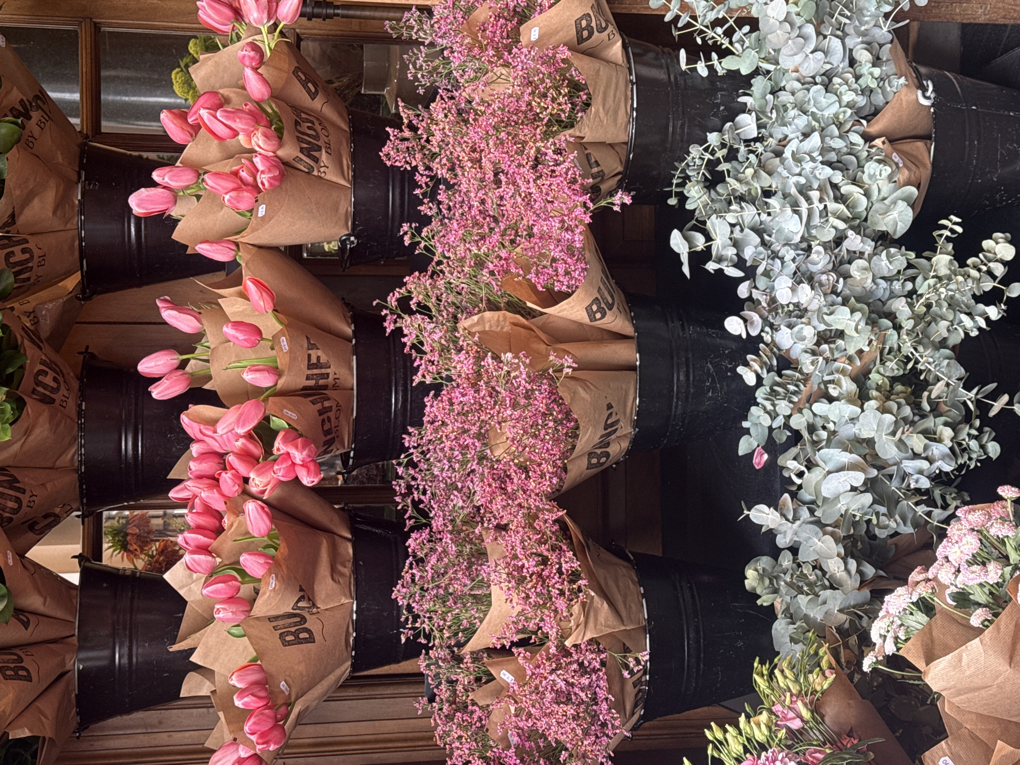 The image depicts several bunches of flowers displayed in black buckets against a wooden wall. The flowers include pink tulips, small pink flowers, and green eucalyptus. The flowers are wrapped in brown paper with the logo "BUNCH BY BLOOM" printed on them. The scene appears to be a flower shop in London, United Kingdom.