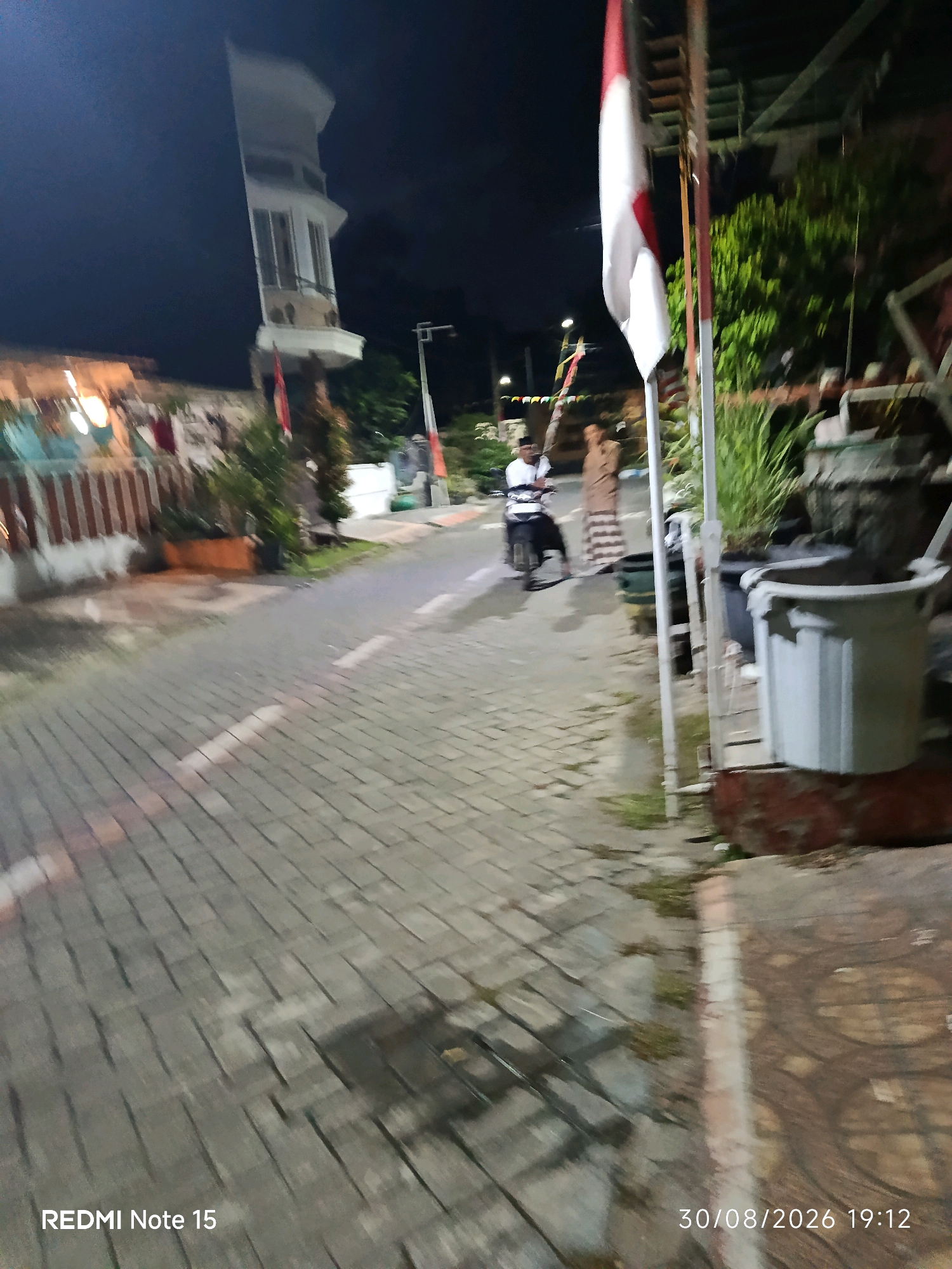 The image depicts a street scene at night in Keboananom, Indonesia. A paved road with brick-like patterns leads through the frame, marked with white dashed lines. On the left side of the road, a building with a white tower structure is visible, with lights illuminating its windows. A red and white Indonesian flag is planted near the tower. Further down the road, a person on a motorcycle, wearing a white shirt and dark pants, is being followed by another individual in a brown outfit. Several potted plants and large containers are arranged on the right side of the road, with one large, light-colored plastic container prominent in the foreground. The overall ambiance suggests a residential or public area at night.