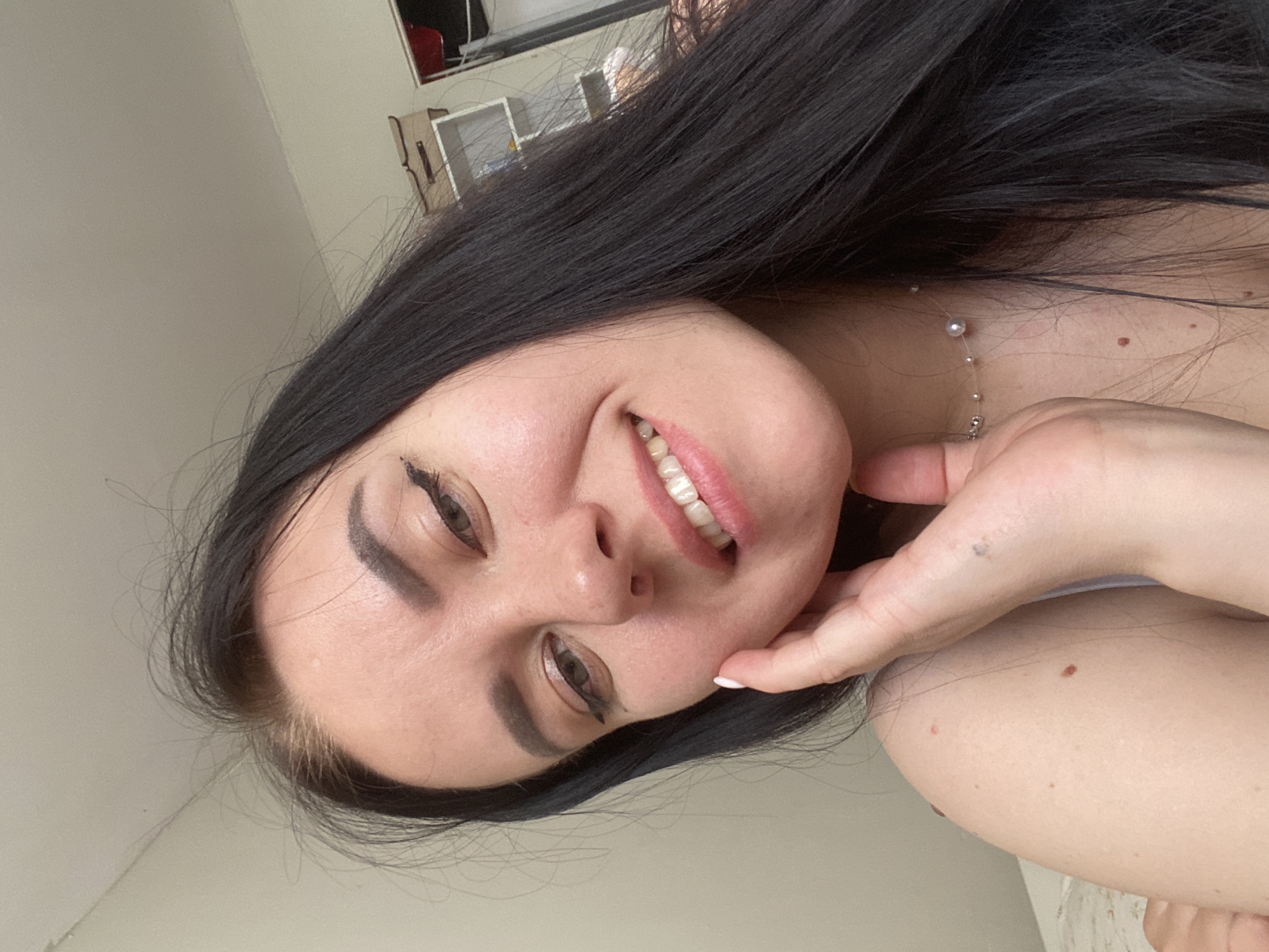 A woman with long black hair smiles while holding her chin. She is wearing a delicate necklace. The background is out of focus but appears to be an interior setting with light-colored walls. The location is not identifiable as Chaikovsky, Russia, based on the visual information provided in the image.