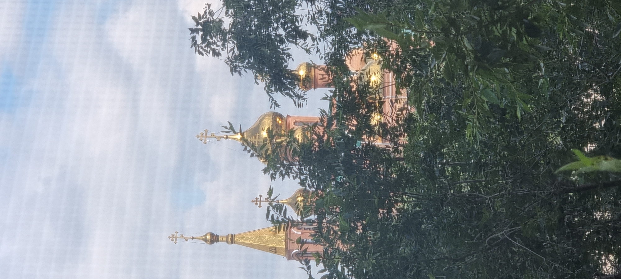 This image captures a view of the Church of the Holy Archangel Michael, a prominent Orthodox Christian church located in the city of Aktobe, Kazakhstan. The church features several golden domes topped with crosses, reflecting the light of the sky, which is a mix of pale blue and white clouds. The foreground is dominated by lush green foliage, with branches and leaves obscuring a significant portion of the church's facade. The building itself appears to be constructed of light-colored brick, with architectural elements that suggest a traditional Orthodox design. The perspective suggests the photo was taken from a slightly elevated position, looking up towards the church through the trees. There are no people visible in the image. The overall impression is one of a religious structure nestled within a natural, leafy environment, partially hidden by the surrounding vegetation.