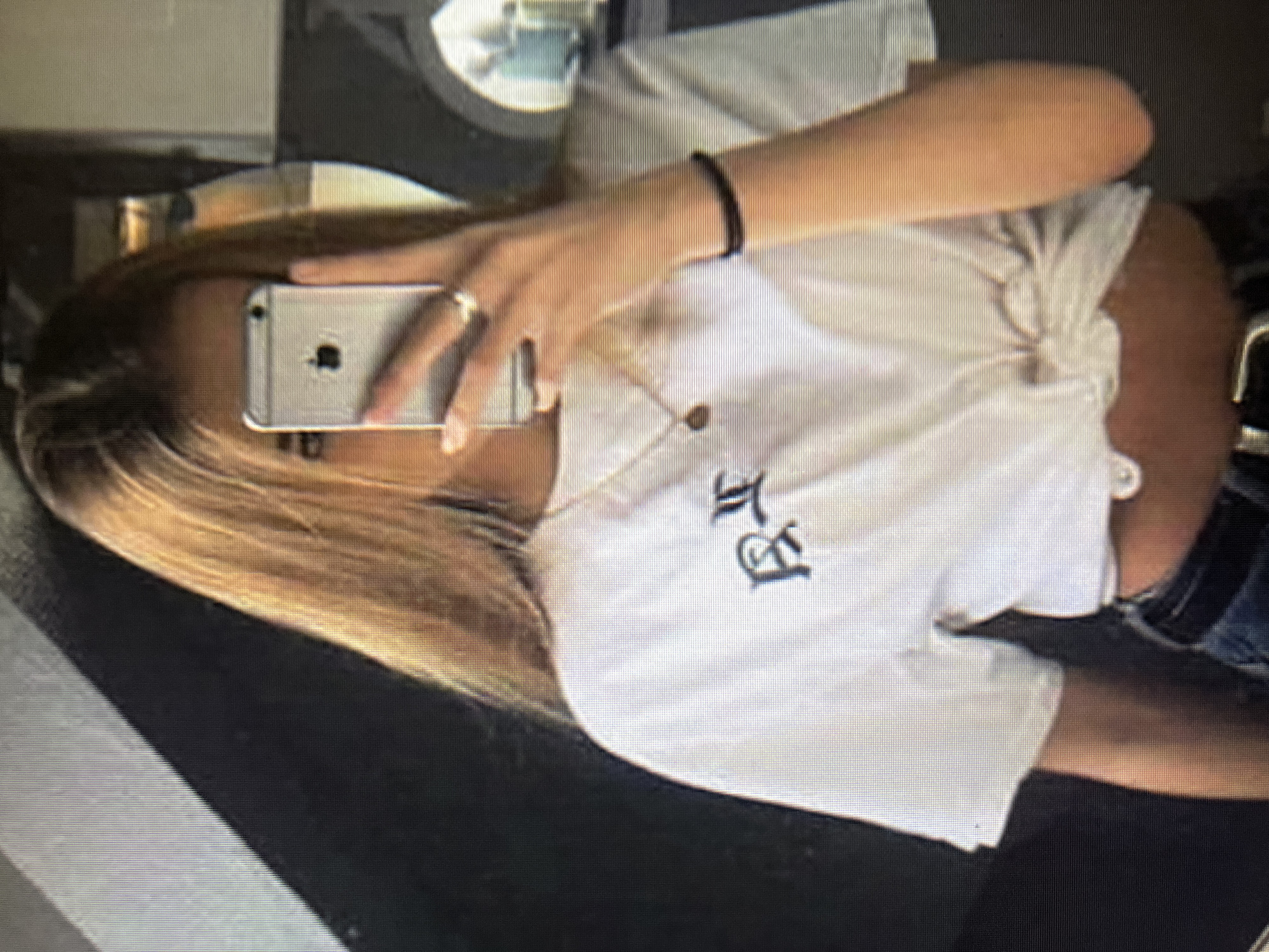 A young woman with long blonde hair is taking a selfie in what appears to be an indoor setting with a dark, textured wall behind her. She is wearing a white t-shirt tied at the waist, revealing a portion of her midriff, and dark denim jeans. On her t-shirt, there is a black graphic design. She is holding a silver smartphone in her right hand to take the photo, and she is wearing a ring on her left ring finger and a black bracelet on her right wrist. A thin gold necklace with a small pendant is visible around her neck. The lighting in the image is somewhat dim, and there are reflections in the background, suggesting a mirror is being used for the selfie. There are no discernible elements that indicate the specific location within the city administration of Qarağandı city, Kazakhstan.