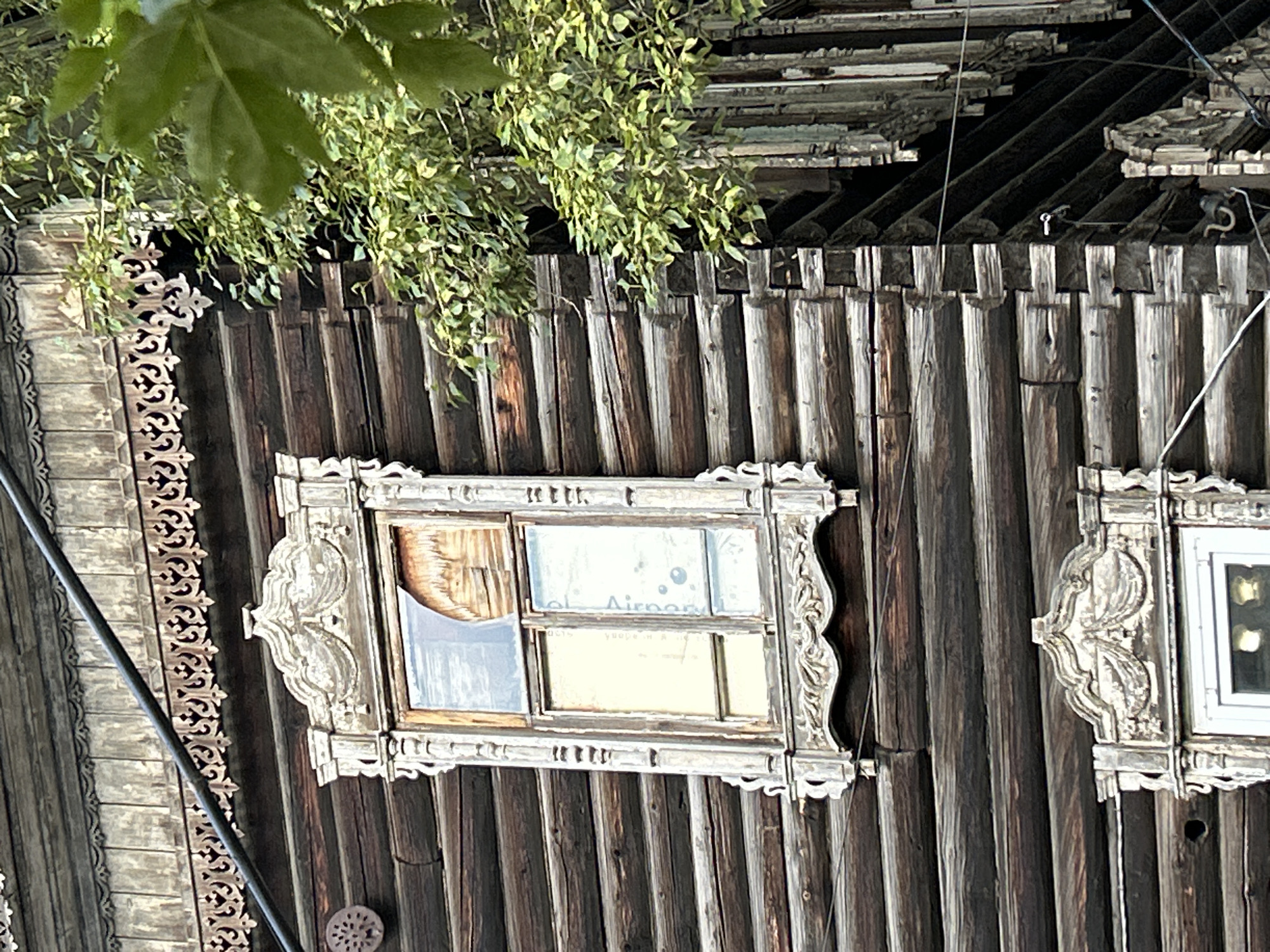 The image shows the side of a weathered, dark wooden log house, characteristic of architecture found in Tomsk, Russia. The house features ornate, carved wooden window surrounds, some with chipped paint, indicating age and exposure to the elements. One window pane is broken, revealing the wooden interior. A tree with green leaves partially obscures the right side of the house. A dark electrical cable runs horizontally across the top of the image. The style of the wooden carvings suggests traditional Russian folk art. The scene is likely a residential area in Tomsk, with the architectural details being a common feature of historical buildings in the city.