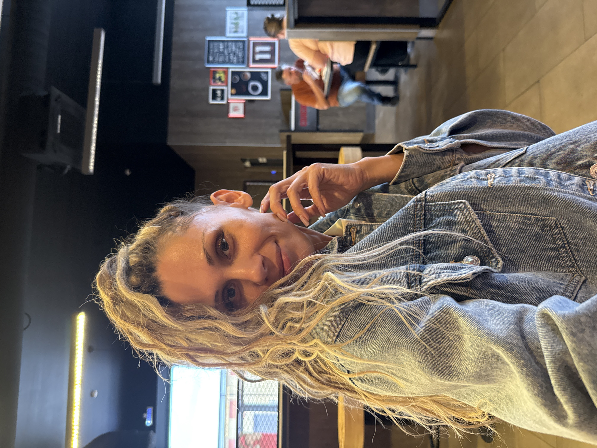 In a casual indoor setting, a woman with blonde, curly hair pulled back in a ponytail and a denim jacket is featured in a close-up shot. She is looking at the camera with a slight smile, her right hand resting near her chin. The background shows a dining area with other patrons seated at tables. The environment appears to be a fast-food restaurant with dark walls, framed pictures, and overhead lighting. The specific location is identified as Ignatovo, Russia.