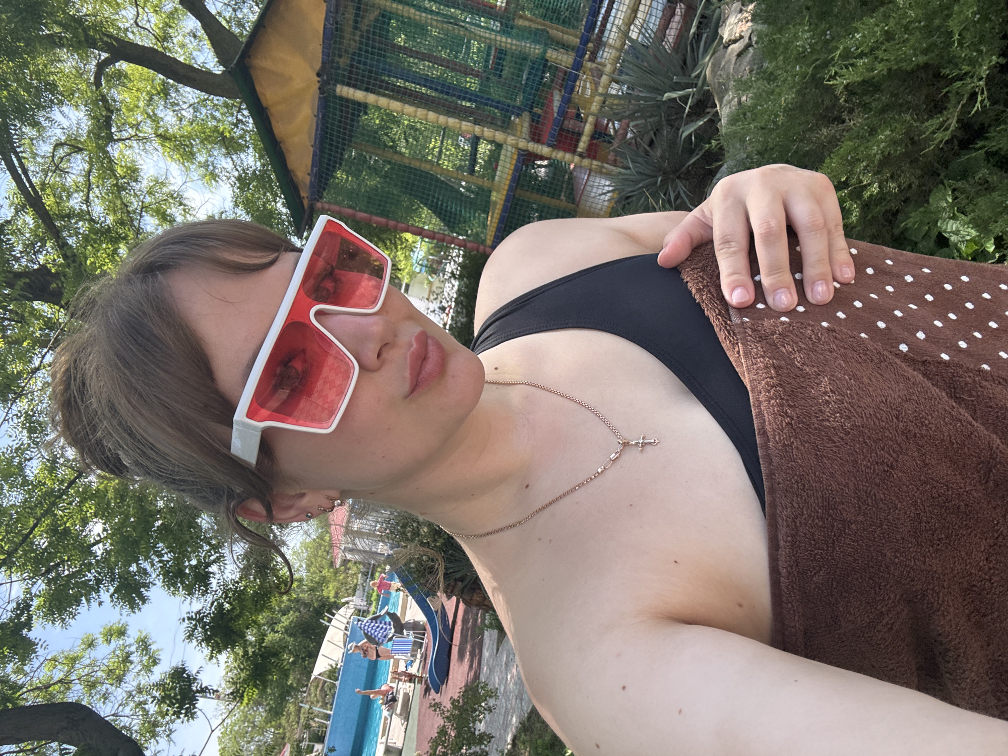 A selfie taken at the AquaLand "U Lukomorya" water park in Yevpatoriya, Ukraine. The subject is a fair-skinned person wearing white-framed red-lensed sunglasses and a black bathing suit top. They have short, light brown hair and are wearing a gold necklace with a cross pendant. A brown towel with white polka dots is draped over their shoulder. The background shows a swimming pool area with people, a slide, and trees. A multi-level play structure with netting is also visible. The sky is visible through the trees, indicating it is daytime.