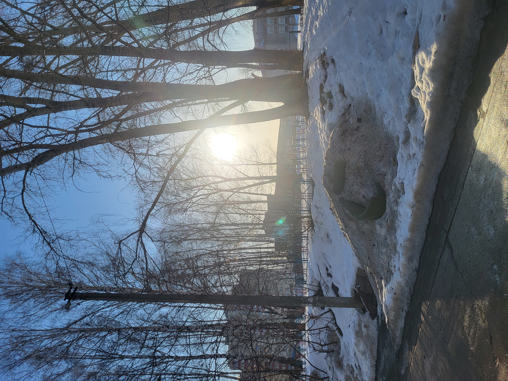 The image captures a street scene in Chaikovsky, Russia, on a bright day. The foreground features a sidewalk, heavily bordered by a mound of snow and ice, indicative of recent winter conditions. Bare tree branches fill much of the frame, silhouetted against a bright, almost blinding sun that dominates the upper part of the image. Through the trees, a glimpse of a building can be seen, appearing to be a multi-story apartment complex, providing a hint of the urban setting. A utility pole, also partially obscured by branches, stands on the left side. The background reveals a playground, further confirming the residential nature of the location. The overall atmosphere is one of late winter or early spring, with the sun's intensity suggesting a warming trend despite the remaining snow.