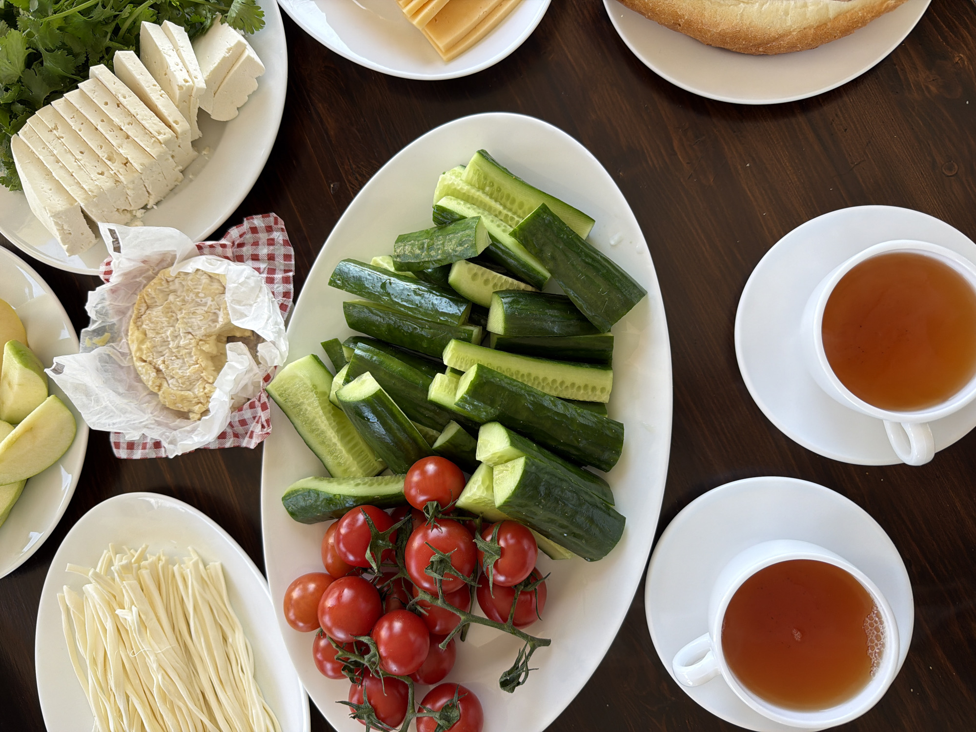 An overhead photograph captures a neatly arranged spread of food and beverages on a dark brown wooden table with a visible grain. The setting suggests a casual breakfast, brunch, or light meal.
Various white ceramic plates and platters are laden with fresh ingredients. In the upper left, a round plate holds several thick slices of white, firm cheese, possibly a type of feta or farmer's cheese, alongside a cluster of fresh green herbs, likely cilantro or parsley. Below this, a small, round soft cheese, possibly Camembert, is partially unwrapped in white paper and sits on a red and white checkered napkin.
A prominent large oval platter in the center features an assortment of chunky green cucumber slices and bright red cherry tomatoes still attached to their vines. To the lower left, another oval plate contains numerous thin, white strands of cheese, resembling string cheese. Sliced green apples are visible on a plate to the far left. Towards the upper center, a plate displays rectangular slices of yellow cheese, while a portion of a crusty baguette or bread roll is seen on a plate in the upper right quadrant.
Two white teacups, each on its matching saucer, are placed on the right side of the table, filled with amber-colored tea. The lighting is bright and even, indicating a daytime scene. No people are visible, and there is no discernible text. The arrangement emphasizes fresh produce and dairy, suggesting a wholesome and inviting meal.