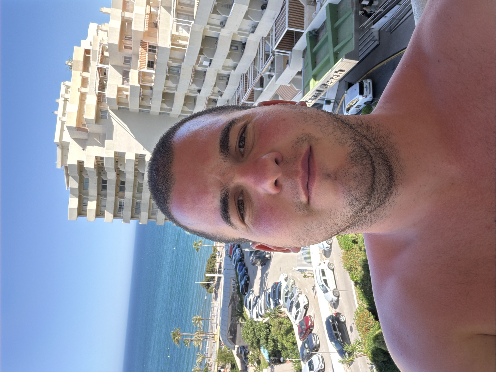 A man with short dark hair and stubble is centered in the frame, facing the camera. He appears to be taking a selfie. Behind him, a tall, multi-story, light-colored building with numerous balconies rises. Further in the background, the blue sea stretches towards the horizon under a clear blue sky. Palm trees line the coast. A road with parked cars runs alongside the beach, and a green area with trees separates the road from the building. The location appears to be the city of Arroyo de la Miel-Benalmádena Costa, Spain, based on the architecture, coastal features, and vegetation.