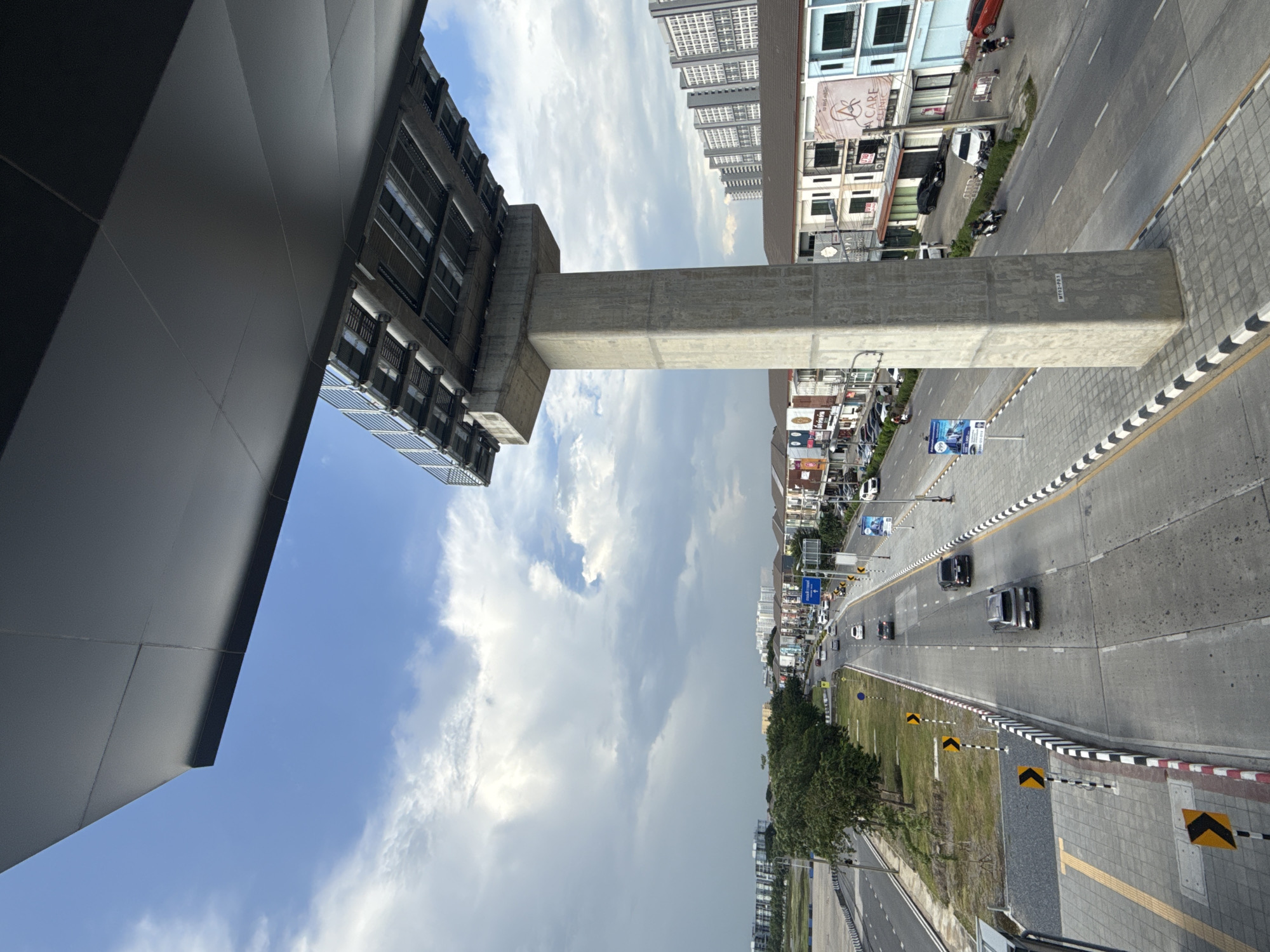An elevated perspective captures a street scene in Pak Kret City Municipality, Thailand, under a partly cloudy sky. A large, dark gray overhang partially obscures the top of the frame, supported by a substantial concrete pillar that stands prominently in the mid-right of the image. The pillar is labeled with the text "MT02-P01".
Below the overhang, a multi-lane road stretches into the distance. On the right side of the road, several buildings with shops and businesses are visible, featuring colorful signage. A white car and a motorcycle are parked in front of one of these buildings. Further down the road, more cars and motorcycles are in motion, indicating moderate traffic.
On the left side of the road, a green median strip separates the lanes from a sidewalk area. This sidewalk features yellow tactile paving. Several yellow and black chevron signs are posted along the roadside, indicating curves or hazards. In the background, a row of trees lines the median, and beyond them, apartment buildings rise against the sky. The overall impression is of a functional urban environment on a day with variable weather.