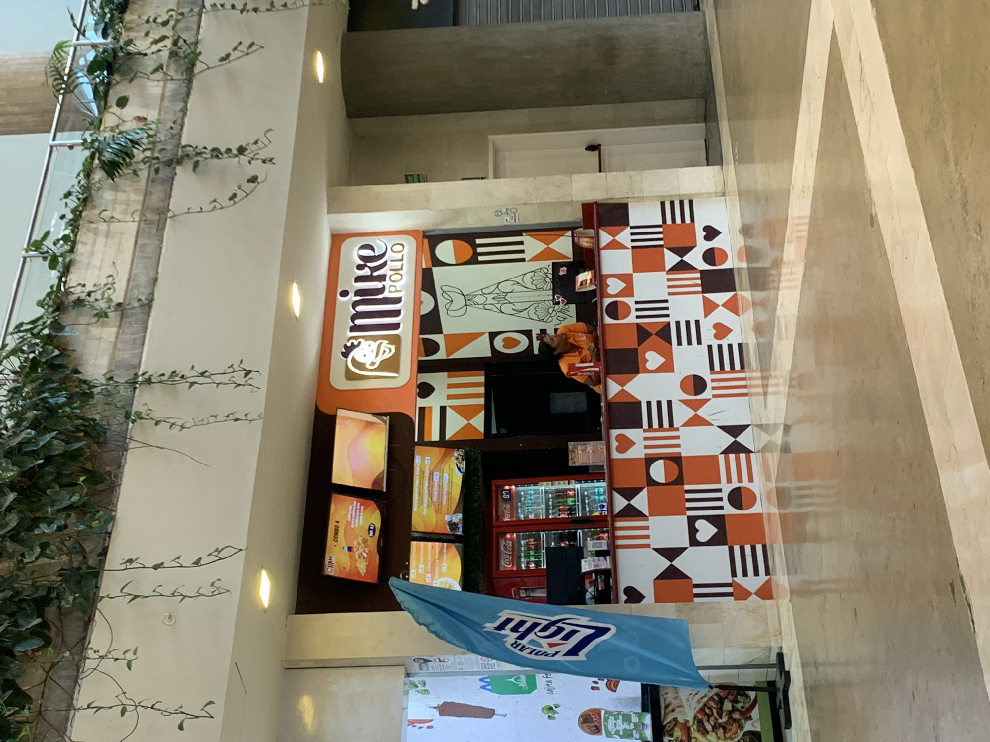 This image captures a vibrant food stall named "Mike Pollo" situated in what appears to be an indoor commercial space, likely a shopping mall or food court, in Pampatar, Venezuela. The stall features a distinct design with its name prominently displayed on an orange-framed sign. Below "Mike" is a cartoon chicken character, and "POLLO" is vertically stacked next to it.
The stall's counter area is adorned with decorative panels featuring a repeating pattern of orange, white, and black geometric shapes including hearts, circles, and stripes. Behind the counter, a staff member in an orange uniform is visible. To the left of the counter, several digital menu screens display food items, one of which clearly reads "COMBO 4". Two large red Coca-Cola refrigerators are also visible, stocked with beverages, and some text on them reads "Coca-Cola" and "87".
In the foreground, a light blue umbrella is partially seen, inverted due to the image orientation, with the text "POLAR Light" (likely a local beer brand) visible. The floor in front of the stall is a beige-tiled surface with a bold white geometric pattern.
To the far left of the image, a light-colored pillar or wall section is covered with lush green vines, adding a touch of natural element to the urban interior. The overall setting appears modern and well-lit by both ambient and artificial lighting. The visible text "Mike Pollo", "Coca-Cola", and "Polar Light" reinforce the commercial nature of the scene and connect to the given location context of Pampatar, Venezuela.