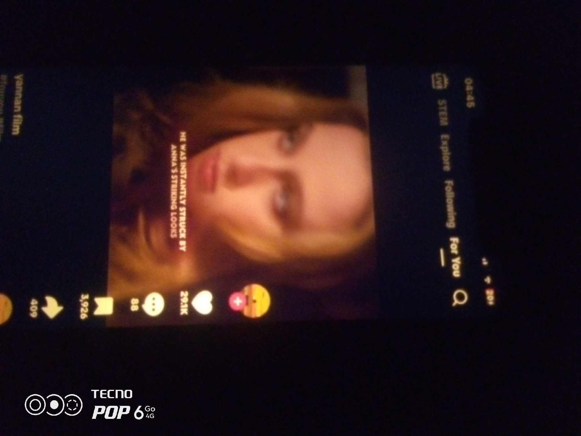 The image displays a dimly lit scene, likely a smartphone screen, showing a short video with overlaid text and icons. A person's face, with blonde hair and light-colored eyes, is centrally featured, looking slightly upwards. Text in white reads, "HE WAS INSTANTLY STRUCK BY ANNA'S STRIKING LOOKS." To the left of the video, text in a green hue reads, "yannan film #tonkikst".
On the right side of the screen, there is a vertical navigation bar with icons and text, including "LIVE," "04:45," "STEM," "Explore," and "Following." Further down, a magnifying glass icon and "For You" are visible.
Along the bottom edge of the video, there are icons representing engagement metrics: a share icon with "409," a bookmark icon with "3,926," a speech bubble icon with "88," and a heart icon with "291K." A pink and yellow circular graphic with a plus sign is also present.
At the very bottom of the image, a brand logo for "TECNO POP 6 4G" is displayed.
The image does not provide any information or visual cues that can be used to determine a location in Howayi, Nigeria, or any other city.