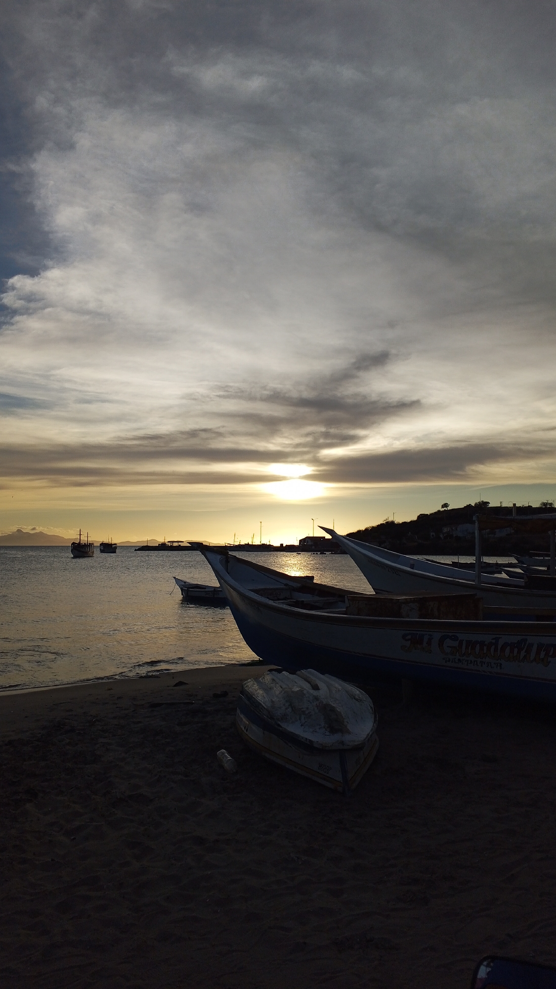 The image depicts a coastal scene at sunset. Several fishing boats are resting on a dark sand beach. The boats are light-colored, with one having the inscription "Mi Guadalupe" visible. In the background, the sun is setting over the water, creating a golden glow. Additional boats are visible on the water, further out from the shore. The sky is filled with clouds. The location is Unknown, Unknown.