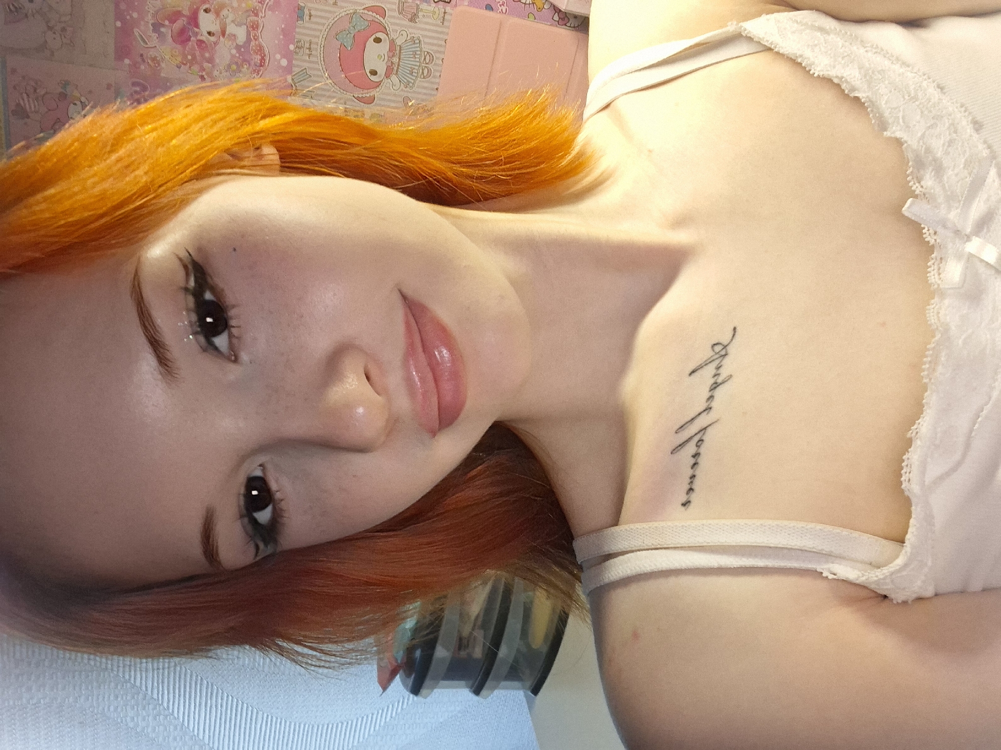 The image shows a young woman with orange hair lying on her side. She has fair skin, freckles, and is wearing eye makeup. She is wearing a white tank top with lace trim. There is a tattoo on her upper chest. The background includes a pink and white wall with cartoon character images. A small item is on the background.
There is no information in the image to determine the location of the image as Chaikovsky, Russia.