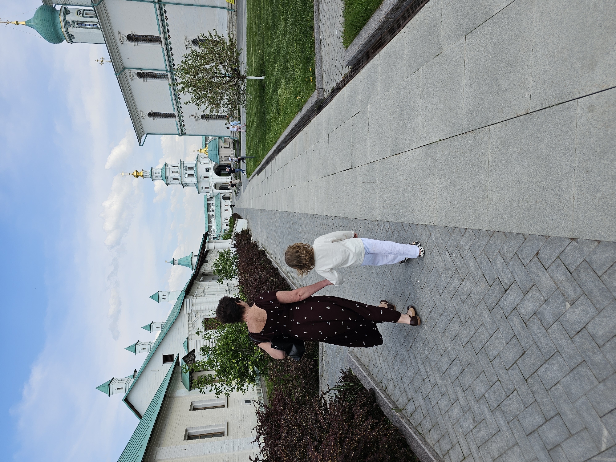 The image depicts a woman and child walking along a paved walkway in Istra, Russia. The walkway is constructed of gray bricks. The woman is wearing a dark, ankle-length dress with a floral pattern. She is holding the child's hand. The child, who has light curly hair, is wearing a white top and white pants. They are positioned on the right side of the image.
To the left of the walkway, there are bushes and the exterior of a white building with green roofs and domes. The sky is partially visible. The background includes several white buildings with green roofs, suggesting a religious complex.