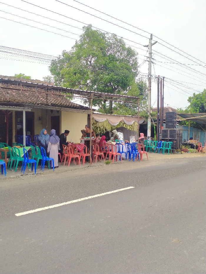 This image depicts an outdoor social gathering or event taking place alongside a paved road in Polokarto, Indonesia, under an overcast sky during daytime. The scene is set in front of a building with a tiled roof, which has an open-air extension covered by a simple metal frame structure. Decorative white and gold fabric drapes are hung along the front of this extension, indicating a special occasion.
Numerous plastic chairs in vibrant red, blue, and green are arranged around several tables on the roadside. People are seen standing, sitting, and interacting. Among them, several women are wearing hijabs. A man in a dark suit and a peci (traditional cap) is visible, engaged with other guests. A woman in patterned attire is seen holding a tray with food, suggesting refreshments are being served.
To the right of the gathering, large black speakers are stacked next to a utility pole, implying that music or public announcements are part of the event. A prominent, large tree with dense green foliage stands in the background, with power lines crisscrossing the sky. The overall impression is that of a communal celebration, possibly a wedding reception or a local gathering, characterized by the informal yet festive setup. No discernible text is visible in the image.