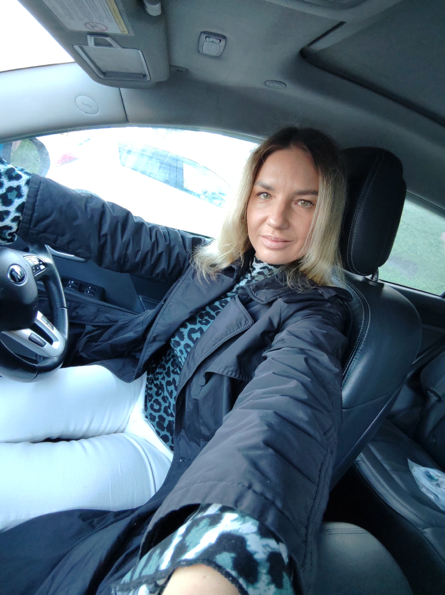 A woman takes a selfie from the driver's seat of a car. She appears to be in her 30s or 40s, with long blonde hair and green eyes, looking directly at the camera with a calm, pleasant expression. She is wearing a dark, possibly black or navy, long coat or jacket, over a teal and black leopard print top, which is visible at her neckline and sleeve cuffs. She is also wearing white pants. Her left hand rests on the steering wheel.
The car's interior is visible, featuring black leather seats, a dark headliner, and a sunroof. A sun visor above the driver's seat has the word "AIRBAG" clearly printed on it. The lighting suggests it is daytime, likely overcast, with diffused light entering through the windows. Through the driver's side window, a blurred outdoor scene with some greenery is visible. A plastic bag rests on the passenger seat. The scene is set in what is identified as Petropavlovsk-Kamchatsky, Russia.