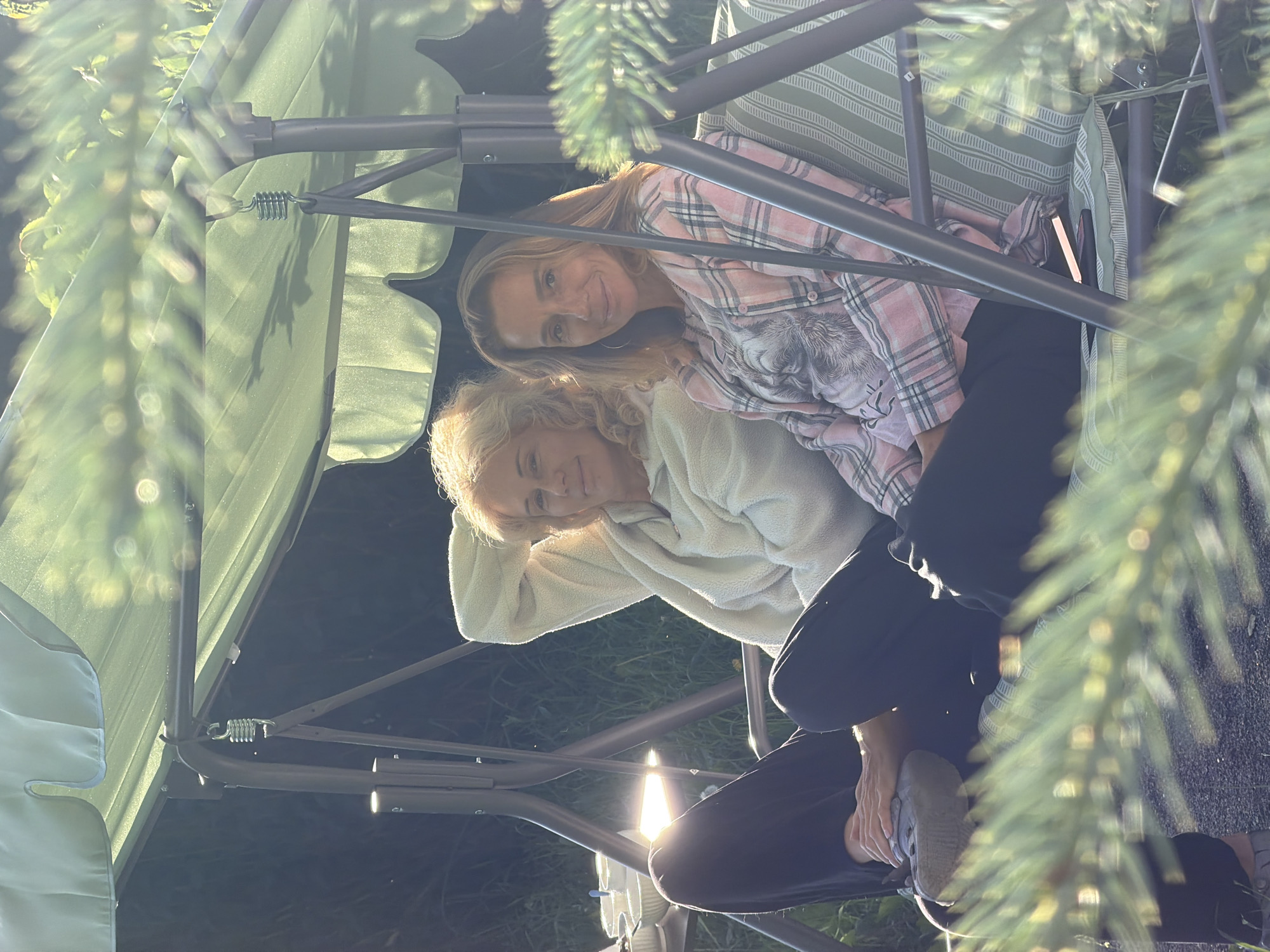 In this photo, two women are sitting on a swing set outdoors. The woman in the foreground is wearing a light-colored, fuzzy sweater and dark pants. She is leaning back, with one arm resting on her head, and her legs are crossed. Her expression appears relaxed. The woman behind her is wearing a pink plaid shirt with a graphic print. She is smiling and looking towards the camera. Sunlight filters through the branches of a tree, casting dappled light on the scene. The background is out of focus, but appears to be a grassy area. The time of day seems to be late afternoon or early evening, given the warm sunlight. The overall mood is one of contentment and casual relaxation. The setting suggests a backyard or garden in Kerro, Russia. There is no visible text in the image.
