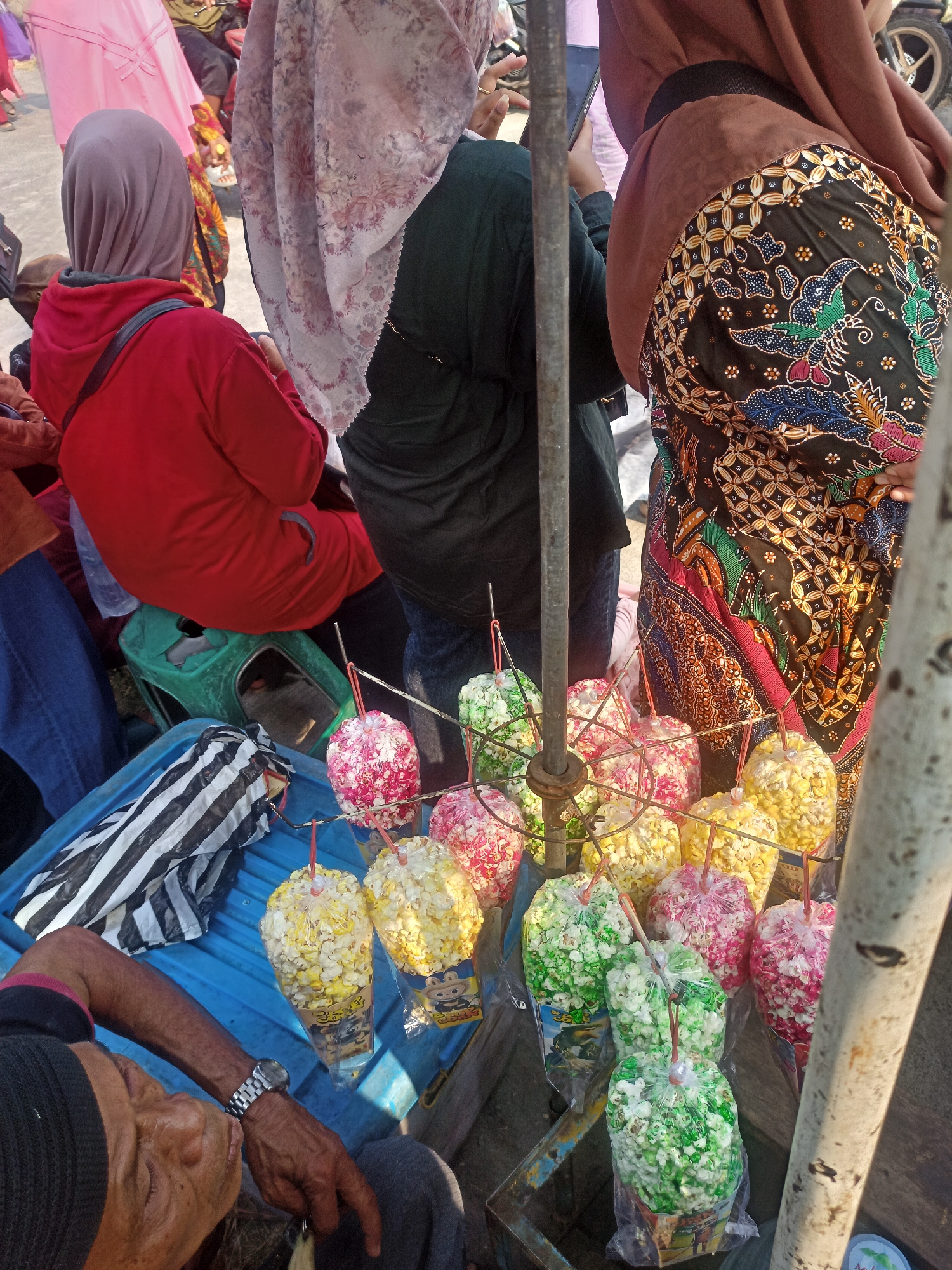 In Gemolong, Indonesia, a street vendor is seen selling colorful bags of popcorn. The popcorn is packaged in clear plastic bags tied with red string, with each bag containing popcorn of a single color: red, yellow, or green. The bags are displayed on a metal rack, some of which have cartoon characters printed on them. In the foreground, an elderly man wearing a black beanie and a watch is partially visible, with his head resting on his arm. Behind him, several individuals are gathered, some wearing headscarves and traditional Indonesian batik clothing. The scene suggests a bustling outdoor market or street event.