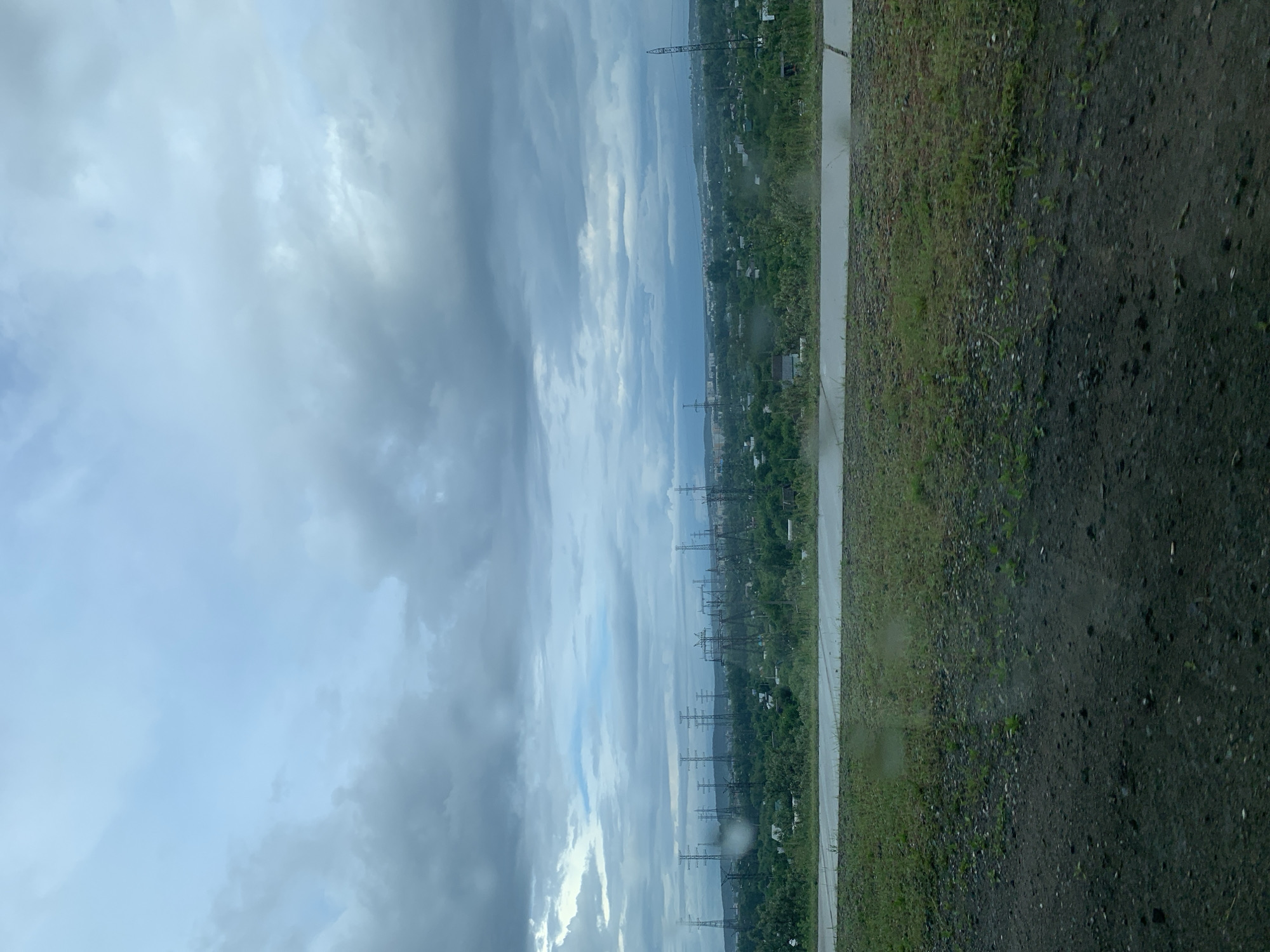 The image captures a wide landscape view in Chaikovsky, Russia, under an overcast sky. The upper portion of the frame is dominated by a vast expanse of heavy, grey and white clouds, suggesting a cloudy day, potentially after rain or during an approaching storm.
Below the sky, the midground features a long, somewhat hazy horizon. A prominent line of numerous tall electricity transmission pylons stretches across the scene, extending into the distance, indicating significant power infrastructure. These pylons are set against a backdrop of dense green vegetation, likely trees and bushes, suggesting a lush, natural or semi-urban environment. A faint, distant line on the horizon could be a body of water or a distant landmass.
In the foreground, the ground is divided into two distinct sections. A light-colored, wet-looking paved surface, possibly a road or a wide path, runs horizontally across the lower part of the image, appearing damp and reflective. Adjacent to this paved area, closer to the viewer, is a darker section of ground composed of dirt and gravel, interspersed with sparse patches of green grass and weeds. The overall dampness of the ground reinforces the impression of recent rainfall.
No specific human activity or event is taking place, and no people are visible. The time of day is daylight, but the heavy cloud cover gives the scene a muted, somewhat somber atmosphere. There is no visible text in the image.