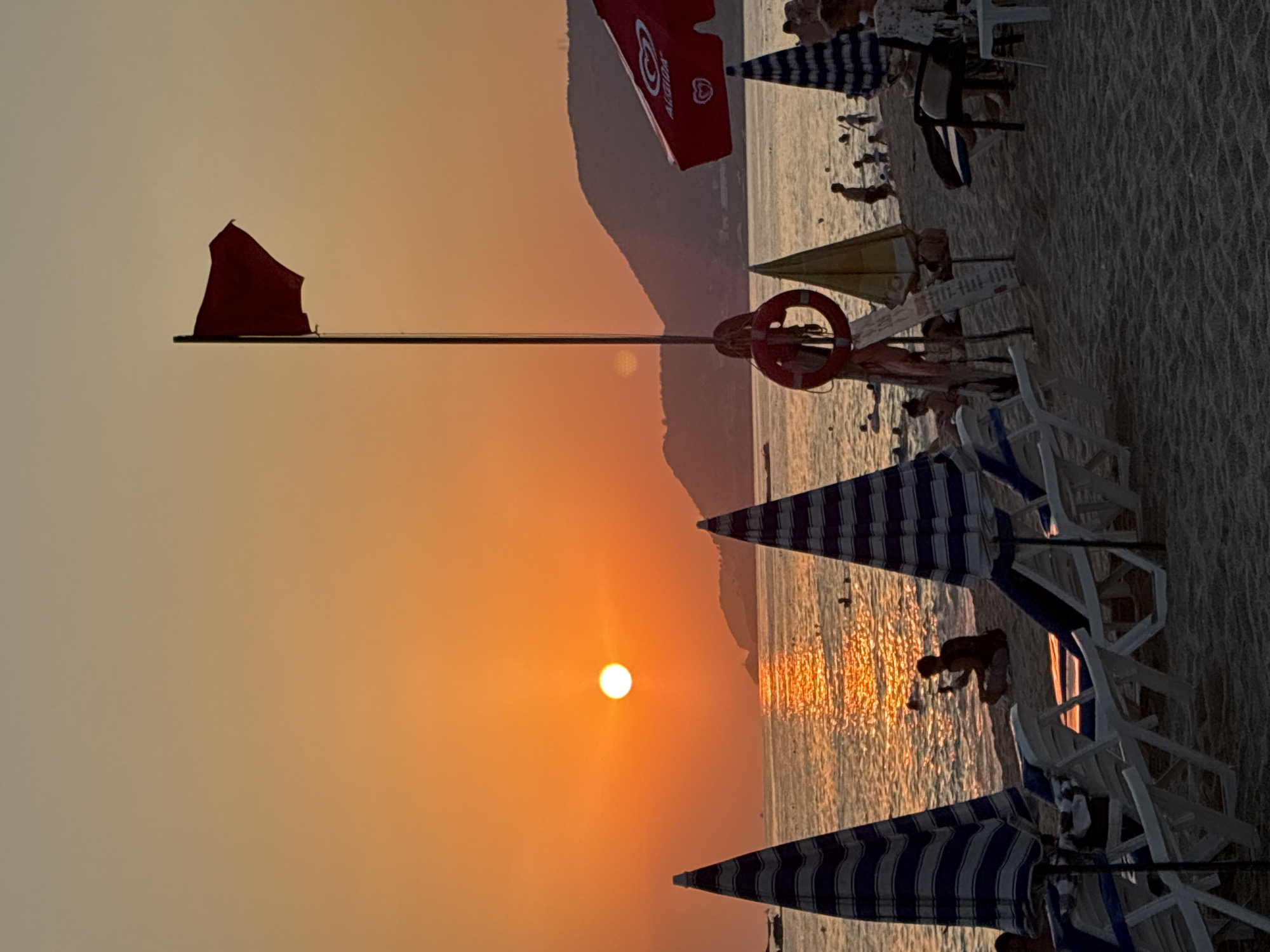 This is a photograph of a beach at sunset in Alanya, Turkey. The sun is low on the horizon, casting a warm orange glow over the sky and sea. The sea is calm with a gentle surf, and there are several people swimming and enjoying the water. The beach is lined with beach chairs and umbrellas, and there are a few people lounging on the sand. A red flag is flying from a pole, indicating caution for swimmers. In the distance, hills and mountains are visible. The overall atmosphere is peaceful and relaxing.