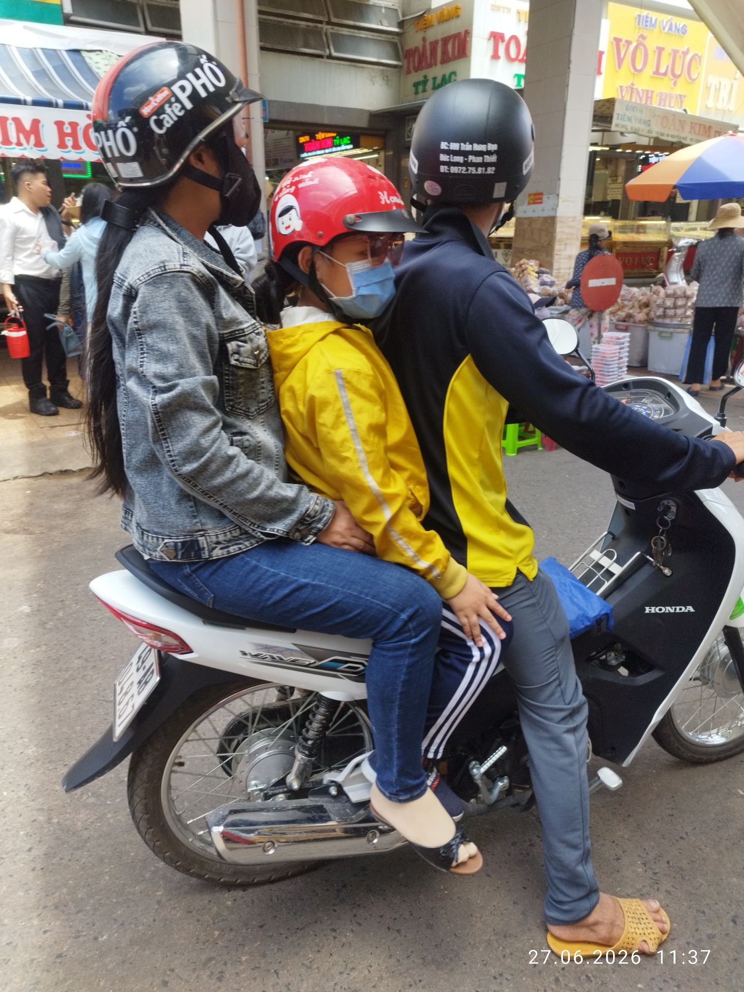 A medium shot captures two individuals on a white Honda motorcycle in Phan Thiết, Vietnam. The person in front, wearing a black and red helmet with "Cafe PHỐ" branding and a denim jacket, is seated behind the driver. The driver, wearing a black helmet, is wearing a black and yellow long-sleeved shirt and gray pants. The motorcycle is positioned on a street with a paved surface. In the background, a market or street vendor area is visible with various stalls and awnings. Several people are seen in the background, including a person in a white shirt and a woman in a straw hat. The overall scene suggests a busy urban environment.
