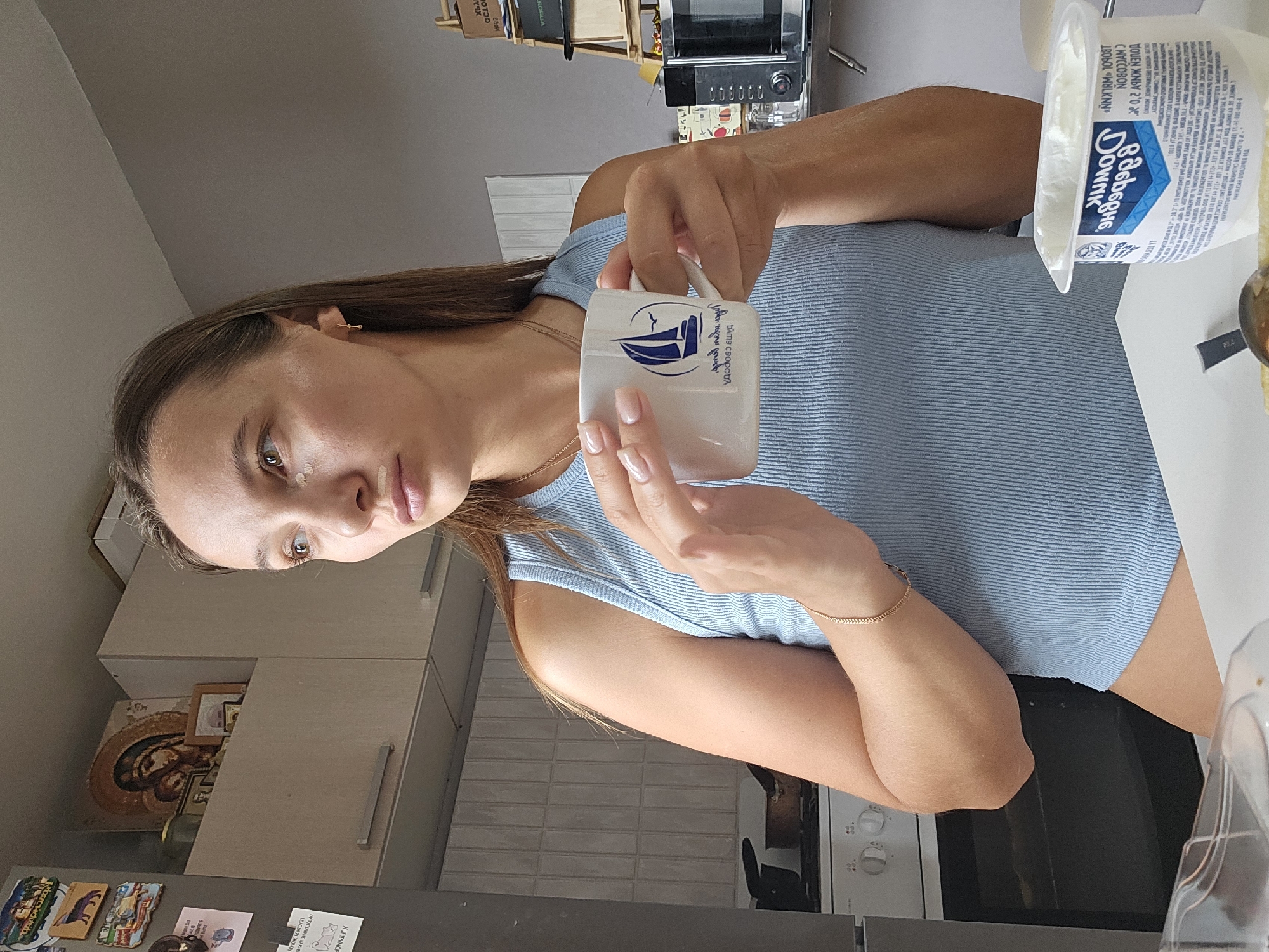 A woman stands in a kitchen, holding a white mug with a blue sailboat logo. She wears a light blue ribbed tank top and a thin gold bracelet. There is a small amount of concealer on her cheeks and nose. The kitchen appears to be in an apartment, with a stove visible in the background. A microwave and shelving unit are also present. The image does not contain information to determine its location within the city of Chaikovsky, Russia.