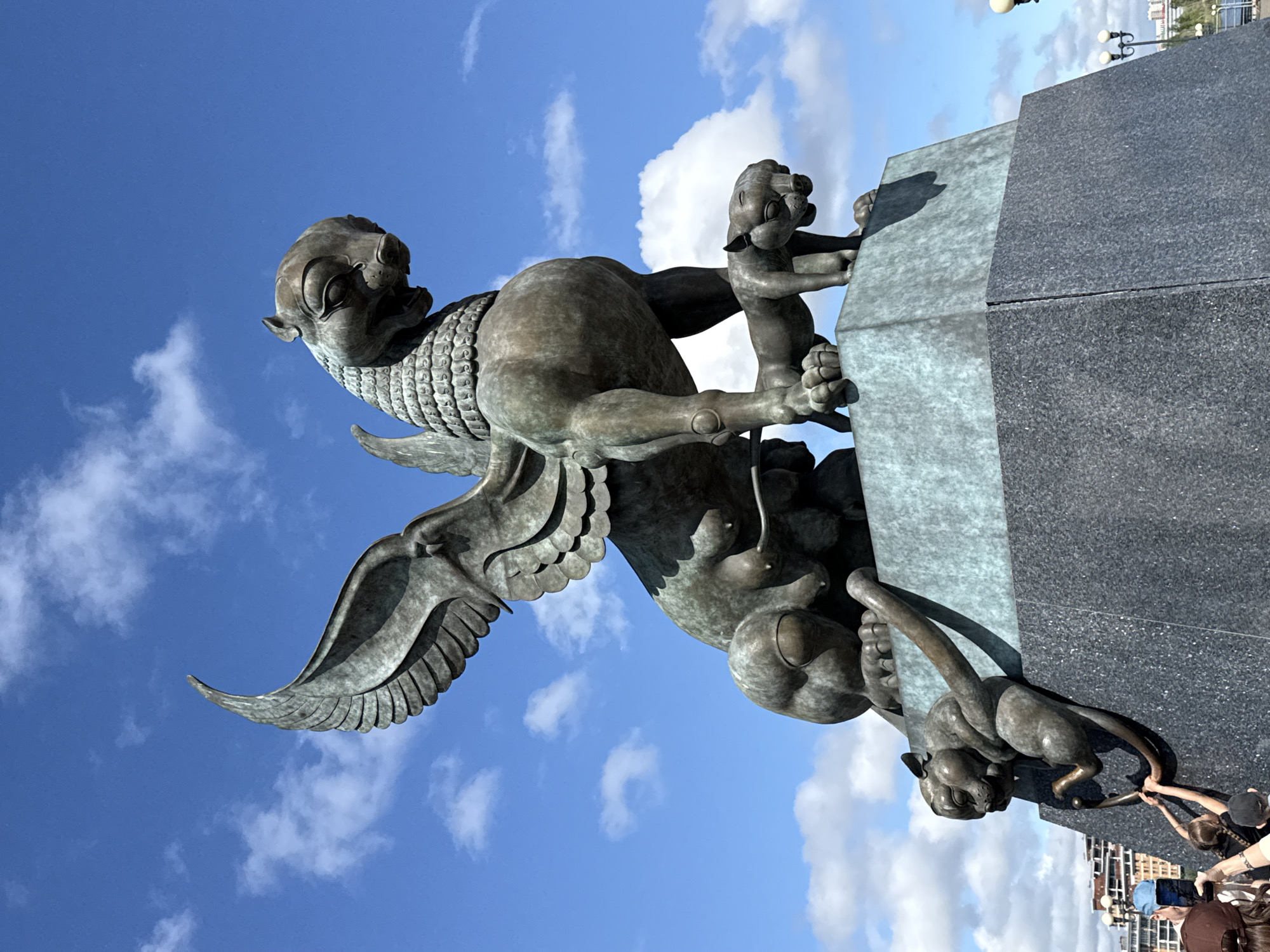 The image displays a large, ornate bronze-colored statue against a clear blue sky with scattered white clouds. The main subject is a prominent winged dragon, or Zilant, a symbol of Kazan, featuring a segmented, scaly body, powerful limbs, and large, detailed wings. Its head has a somewhat feline or draconic appearance. The statue is mounted on a dark, polished stone pedestal, likely granite.
Clinging to the main statue and its base are several smaller, similar mythical creatures, resembling young dragons or griffins. One small creature is depicted climbing onto the pedestal from the main body, while another is positioned lower down on the base.
At the very bottom right of the image, several people are visible, gathered around the base of the statue. They appear to be interacting with the smaller figures, with some reaching out to touch them. Their presence suggests the monument is a public attraction.
The scene takes place outdoors on a bright, sunny day, indicated by the vibrant sky and clear lighting. A traditional lamppost with two lights and distant buildings can be seen in the upper right background, suggesting an urban or park setting. The overall impression is one of a significant public monument being observed and engaged with by visitors in Kazan, Russia. There is no visible text.