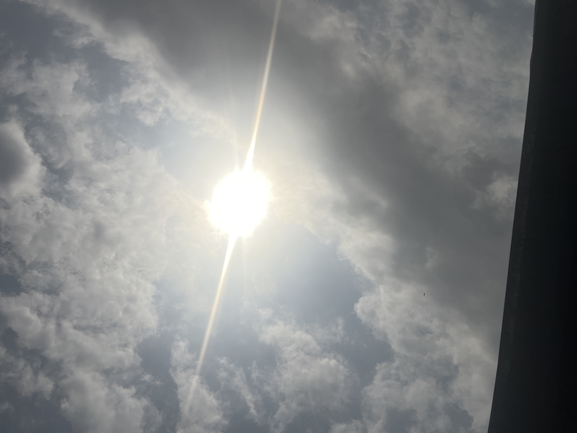 The image presents an upward view of the sky in Ikot Ekpene, Nigeria. A brilliant sun is centrally located, emitting strong horizontal lens flare across the frame. The sky is largely covered by a heterogeneous mass of white and grey cumulus clouds. A dark, indistinct horizontal silhouette, consistent with the upper edge of a building or structure, occupies the lower portion of the image. A single, small, dark, avian-like object is visible in the upper-left quadrant against the cloud background. No human subjects or discernible interactions are present.