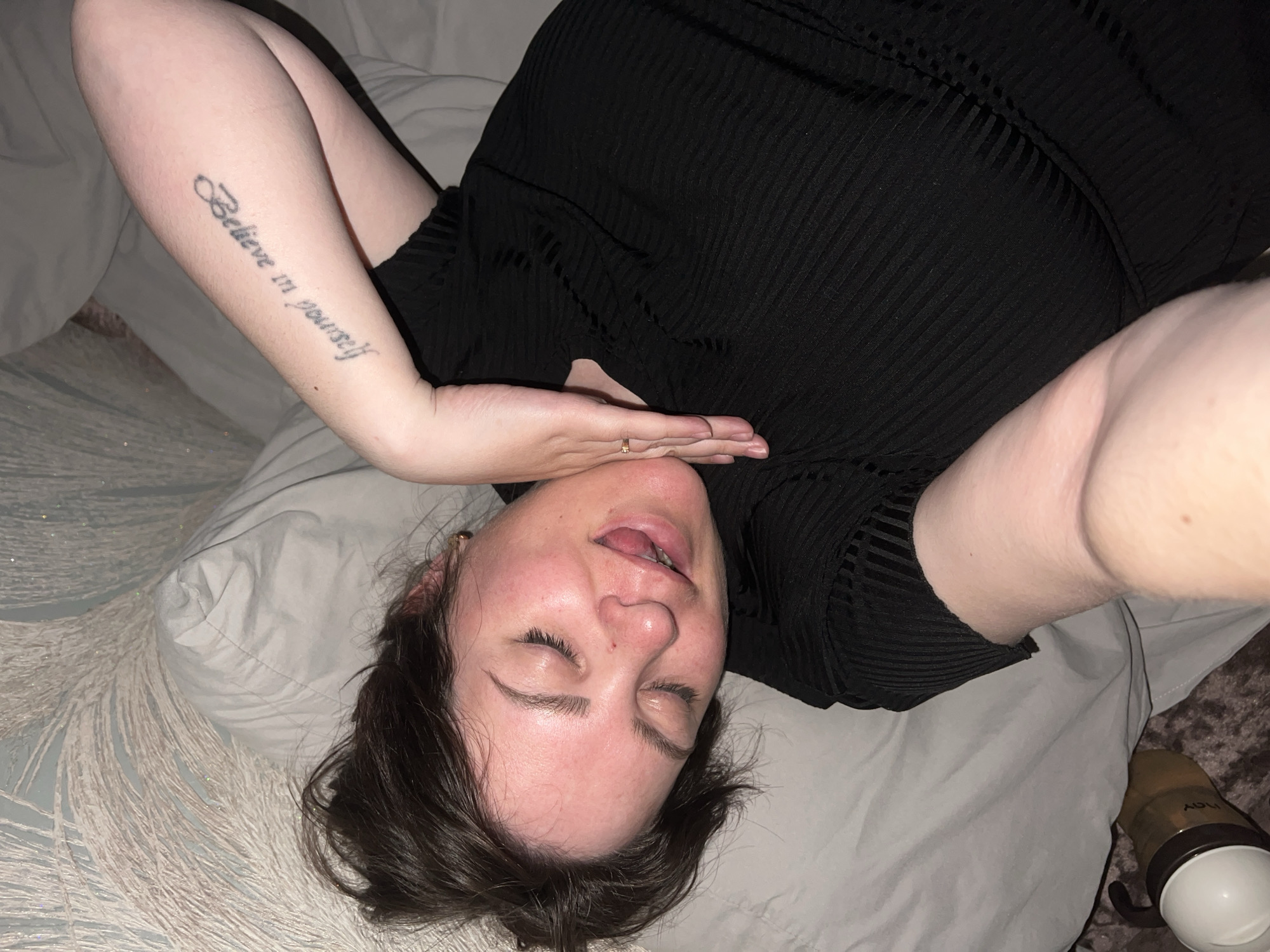A fair-skinned woman with brown hair is lying on her back on a light-colored bed with a textured pattern. She is wearing a black ribbed top. Her eyes are closed and her tongue is slightly sticking out. Her left arm is raised, revealing a tattoo on her inner forearm that reads "Believe in yourself". She is wearing a ring on her left hand. A bottle is visible in the lower right corner of the image. The location is unspecified and not evident from the provided image.