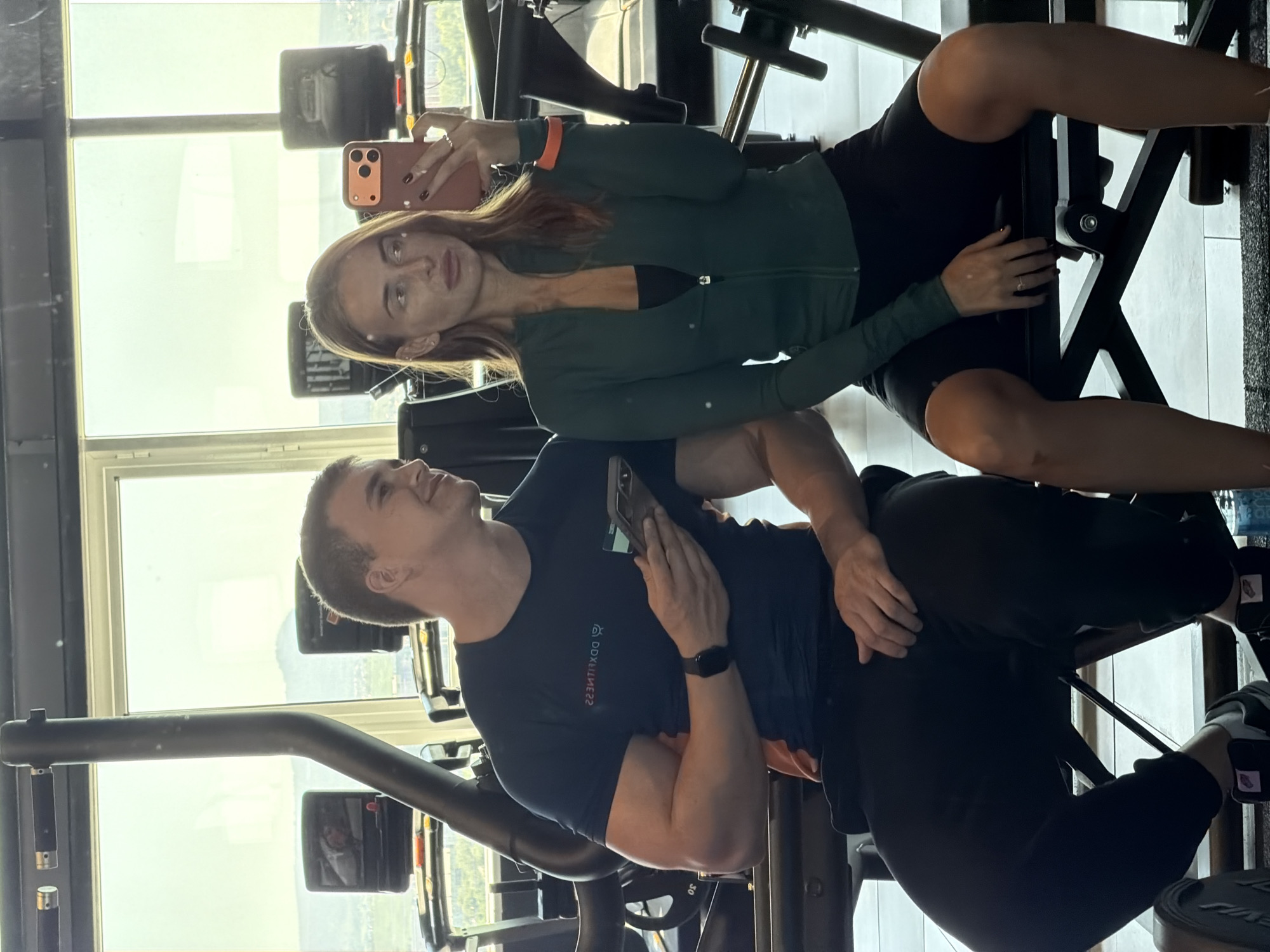 This media file captures two individuals, a man and a woman, inside what appears to be a modern gym with large windows overlooking an outdoor scene. The image is oriented sideways, with the subjects appearing as if lying down or reclined, but they are likely seated on gym equipment.
The woman is positioned at the top of the frame. She has long, light brown hair and is wearing a dark green athletic zip-up jacket over a black sports bra, and black athletic shorts. She is holding a coral-colored smartphone with a distinctive camera module, seemingly taking a selfie. A silver ring is visible on her left hand's ring finger, and she wears an orange band on her right wrist. Her expression is neutral as she looks towards her phone.
The man is positioned below the woman. He has short, light brown hair and is wearing a dark blue t-shirt with white text that reads "DDXFITNESS" horizontally across the chest. He also wears black athletic pants and a black smartwatch on his left wrist. He is holding a dark-colored smartphone in his left hand, resting it on his stomach. He is looking upwards and to his left, with a relaxed, smiling expression.
Both individuals are seated on dark-colored gym equipment, suggesting they are taking a break or posing within the workout facility. The large windows in the background indicate a high-rise location or a gym with a scenic view, allowing ample natural light to illuminate the scene. The bright light outside suggests it is daytime. The overall mood appears casual and relaxed.