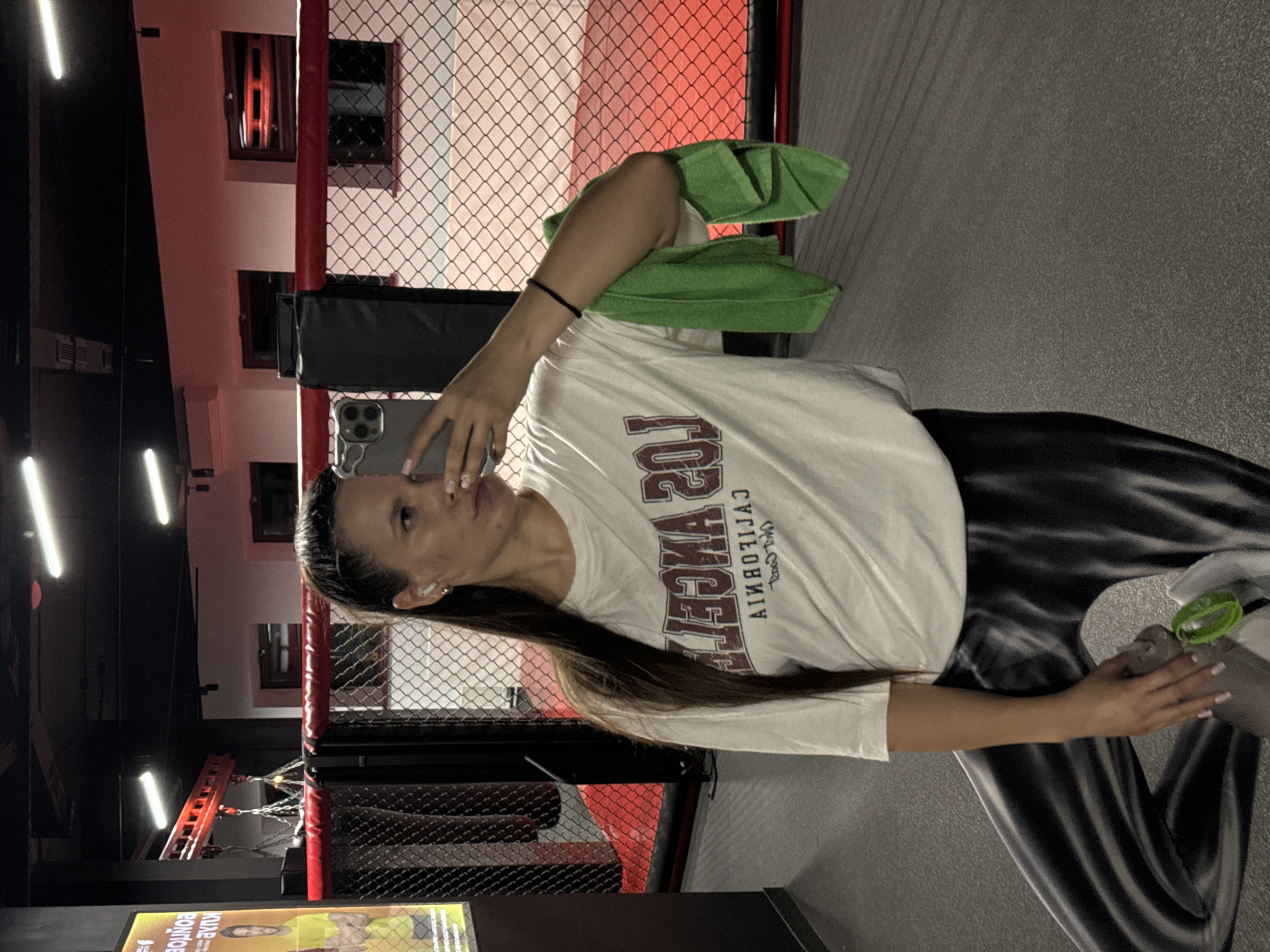 A woman is taking a selfie inside what appears to be a mixed martial arts (MMA) training facility. The woman is holding her phone in her left hand and a green towel is draped over her left shoulder. In her right hand, she holds a water bottle. She is wearing a white t-shirt with "LOS ANGELES CALIFORNIA" printed on it and dark, shiny leggings. Behind her is the chain-link cage of an MMA ring, with red padding on the support beams. There is a gray rubber floor in the training area. Overhead lighting is visible. The location is an MMA gym, the city of Vniissok, Russia cannot be determined.