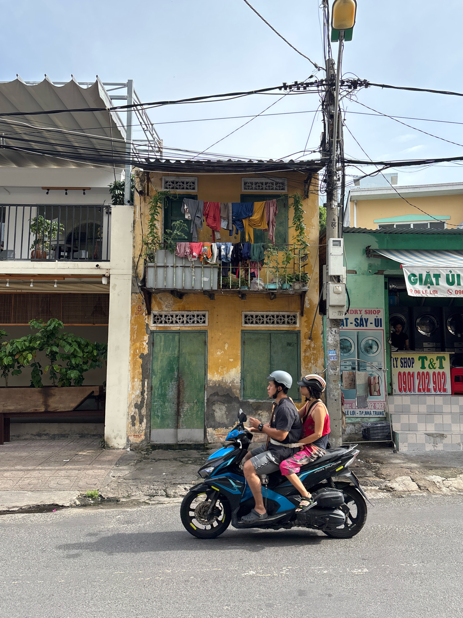 The image depicts a street scene in Nha Trang, Vietnam, likely a residential or mixed-use area. In the foreground, a blue and black motorcycle with two riders, a man and a woman, is moving along an asphalt road. Both riders are wearing helmets. The man is driving, wearing a dark t-shirt and gray shorts. The woman is seated behind him, wearing a red tank top and patterned shorts.
Behind the motorcycle, a yellow building with weathered paint is visible, featuring a balcony with clothes hanging out to dry. The balcony has a metal railing and is adorned with green plants. Below the balcony, there are two green doors. To the right of the yellow building is a green-fronted establishment with a striped awning. Signage above the awning reads "GIẶT ỦI" (laundry) and lists a phone number and address. Inside this establishment, washing machines are visible, and a person is standing behind a counter.
Further to the left, a modern building with a balcony railing and potted plants is present. The street is lined with utility poles carrying numerous electrical wires. The overall atmosphere suggests a typical day in a Vietnamese city, with everyday activities like laundry and transportation by motorbike taking place. The buildings exhibit a mix of older, traditional architecture and newer constructions. The presence of laundry hanging out to dry is a common sight in many urban areas in Vietnam. The context of "TP. NHA TRANG" on the signage in the image confirms the location as Nha Trang.