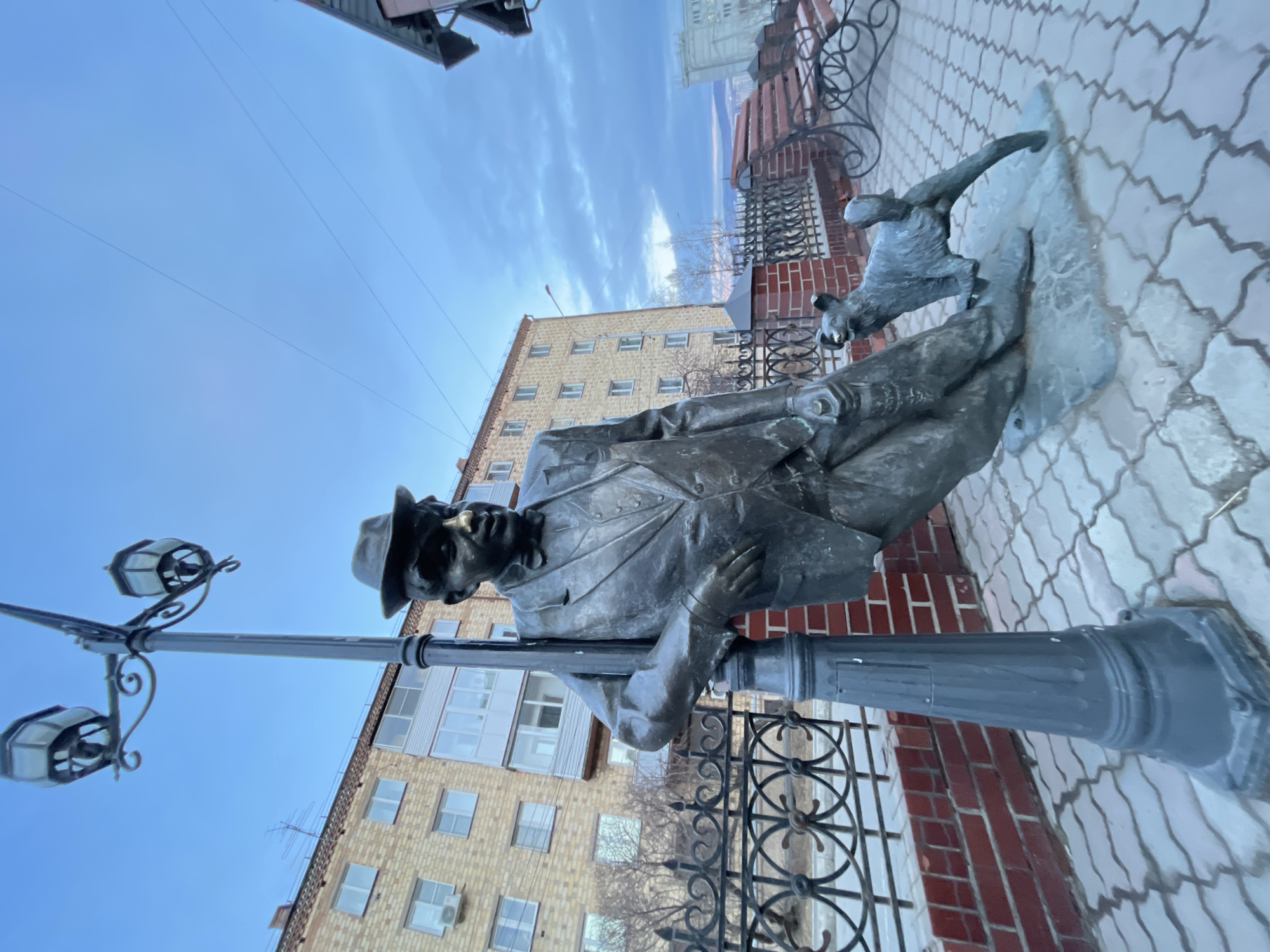 This image captures a bronze statue of a man and a dog situated in an urban environment in Krasnoyarsk, Russia. The primary subject is a full-body statue of a man, depicted leaning casually against a dark, ornate street lamppost on the right side of the frame. He is dressed in a suit, a bow tie, and a fedora hat, with a notable mustache and a contemplative expression. At his feet, a smaller bronze statue of a dog, or possibly a fox, is depicted looking upwards.
The lamppost itself is decorative, featuring two traditional lanterns. The ground beneath the statues is paved with light-colored, interlocking paver stones arranged in a distinct pattern. In the background, several multi-story buildings are visible, including one prominent structure constructed of light brown brick with numerous windows, and another with lighter-colored facades. An ornamental wrought-iron fence separates the statues from the background buildings.
The scene is bathed in natural light under a clear blue sky with scattered clouds, indicating it is daytime. No specific activity or event is taking place beyond the static presence of the public art installation. There is faint, illegible text on a distant building in the background. The overall setting is that of a city street or public square.