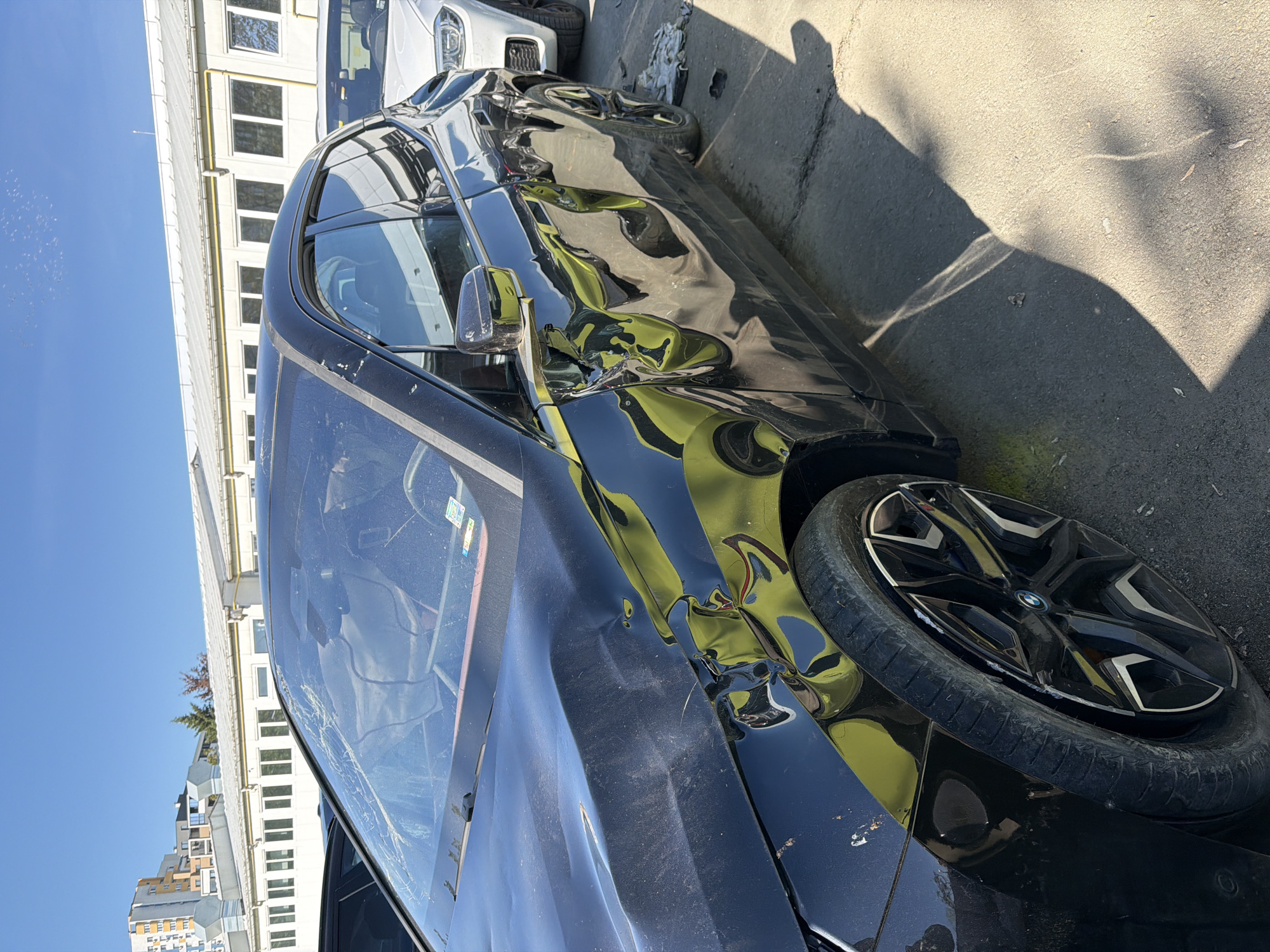 The image shows a black sedan, heavily damaged on the front passenger side. The car is parked on an asphalt surface, likely a street or parking area. The front passenger tire is flat. The side panels of the car are heavily dented. The windshield is cracked. In the background, there's a multi-story building with windows and blue sky is visible in the background. Based on visual characteristics of the multi-story building and the cracked glass on the window, the car incident may have occurred in Burgas, Bulgaria.
