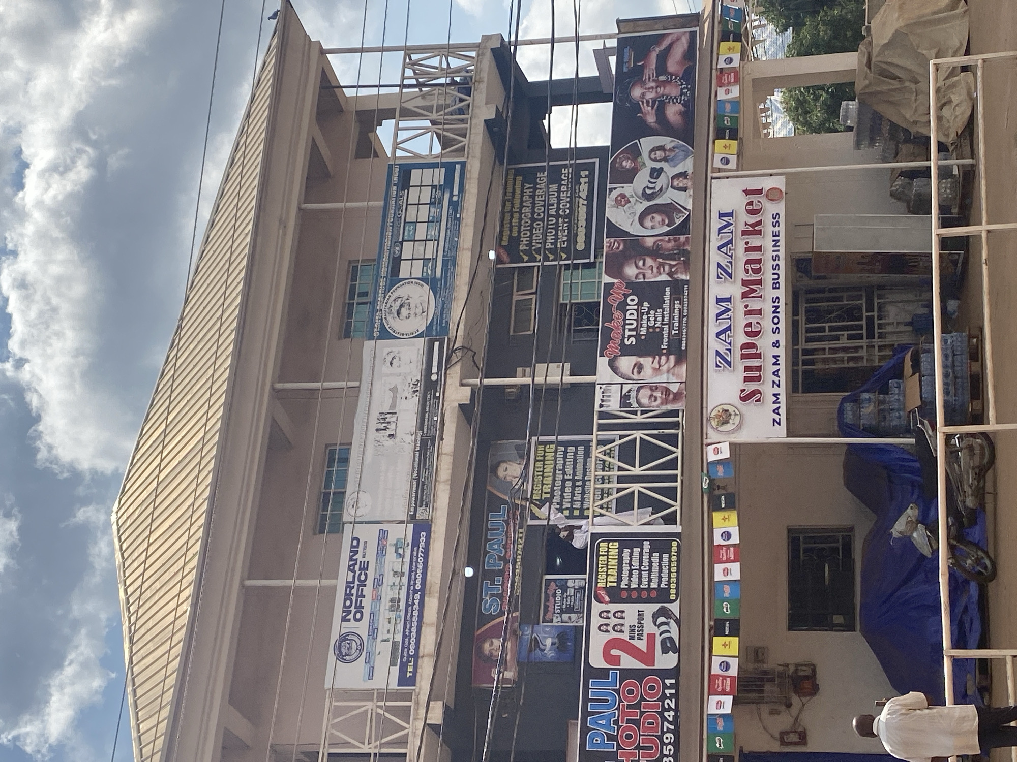 This is a full-shot, eye-level exterior view of a multi-story building in Katambe, Nigeria, under a cloudy sky. The building has a light brown metal roof and appears to be a commercial establishment. Various signs are visible on the facade, advertising services such as "NORLAND OFFICE," "ST. PAUL," "MAKE-UP STUDIO," "ZAM ZAM SuperMarket," and "REGISTER FOR TRAINING" in Photography, Video Editing, Photo Album, and Event Coverage. A man in a light-colored shirt and dark pants is standing on the left, facing the building and holding a phone up, possibly taking a picture. In front of the building, several motorcycles are covered with blue tarps, and another motorcycle is partially visible on the right. Pallets of bottled water are stacked on the right side of the image. Decorative flags are strung across the front of the building above the ground floor shops.