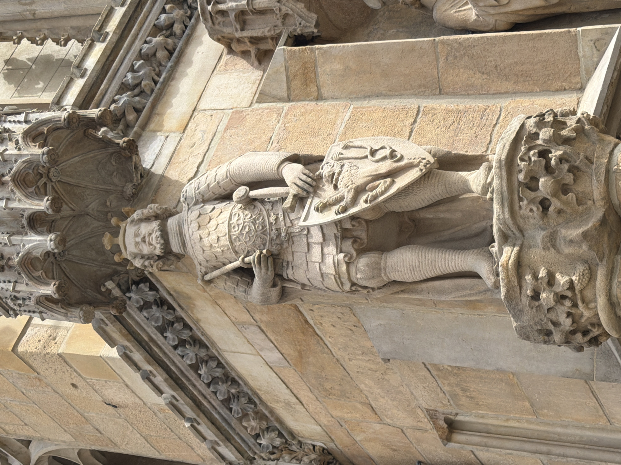 A stone statue of a warrior, likely a king or knight, stands prominently on the corner of a building made of tan stone blocks. The warrior is depicted wearing a crown, armor, and a cape, and holds a sword in his right hand and a shield in his left. The shield features a relief of a lion. The statue is positioned on a pedestal decorated with intricate carvings. The building behind the statue exhibits Gothic architectural elements, including ornate stonework and pointed arches. The lighting suggests it is daytime, with strong shadows indicating the sun is casting light from the right. Another partial statue of a robed figure is visible to the right of the main warrior statue.