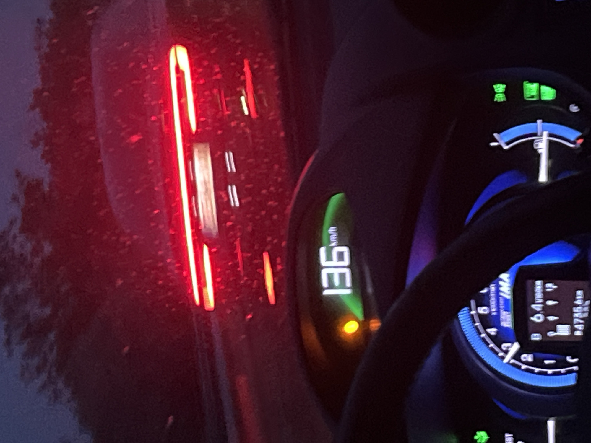 The image captures a nighttime view from inside a vehicle, looking forward at the dashboard and the rear of a dark sedan on a wet road in Gorno Varshilo, Bulgaria. The vehicle's speed is indicated as 136 km/h on a green digital display on the upper dashboard. An illuminated amber warning light is present to the left of the speed display. The lower dashboard features a blue arc gauge, which appears to be a tachometer or hybrid system indicator, with a needle pointing to approximately 2-3 units. A central digital screen displays "IMA," followed by "B 6.4 l/100km" (fuel consumption) and "P 4785.6 km" (odometer or trip meter). To the right, green indicator lights are visible, including a headlight icon and another rectangular status light. The fuel gauge, depicted as a vertical bar, is mostly full.
The dark-colored sedan ahead features distinct horizontal LED taillights emitting bright red light, with two main strips on each side. Smaller lights illuminate the indistinct license plate area. Reflective moisture droplets, indicative of rain or a wet road surface, are scattered across the lead car's rear and the intervening air, likely observed through a rain-splashed windshield. Dark, indistinct foliage lines the roadside under a dark sky, suggesting a rural or semi-rural setting.