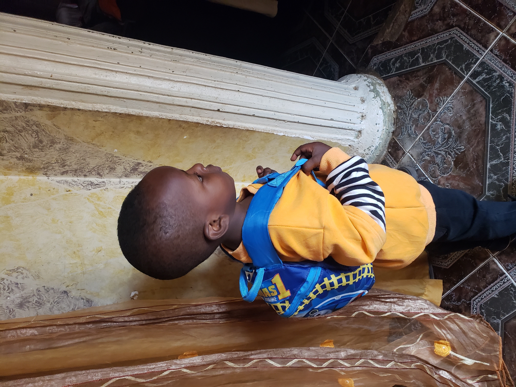 In Sabon Tasha, Nigeria, a young child, viewed from the side, stands near a pillar and a draped curtain. The child is wearing a bright orange long-sleeved shirt with black and white striped cuffs, and black trousers. A blue backpack with yellow accents and a cartoon character is worn on the child's back. The background features a textured yellow wall, a decorative pillar, a translucent brown curtain with embroidered yellow floral patterns, and a patterned tiled floor. The lighting suggests an indoor environment.