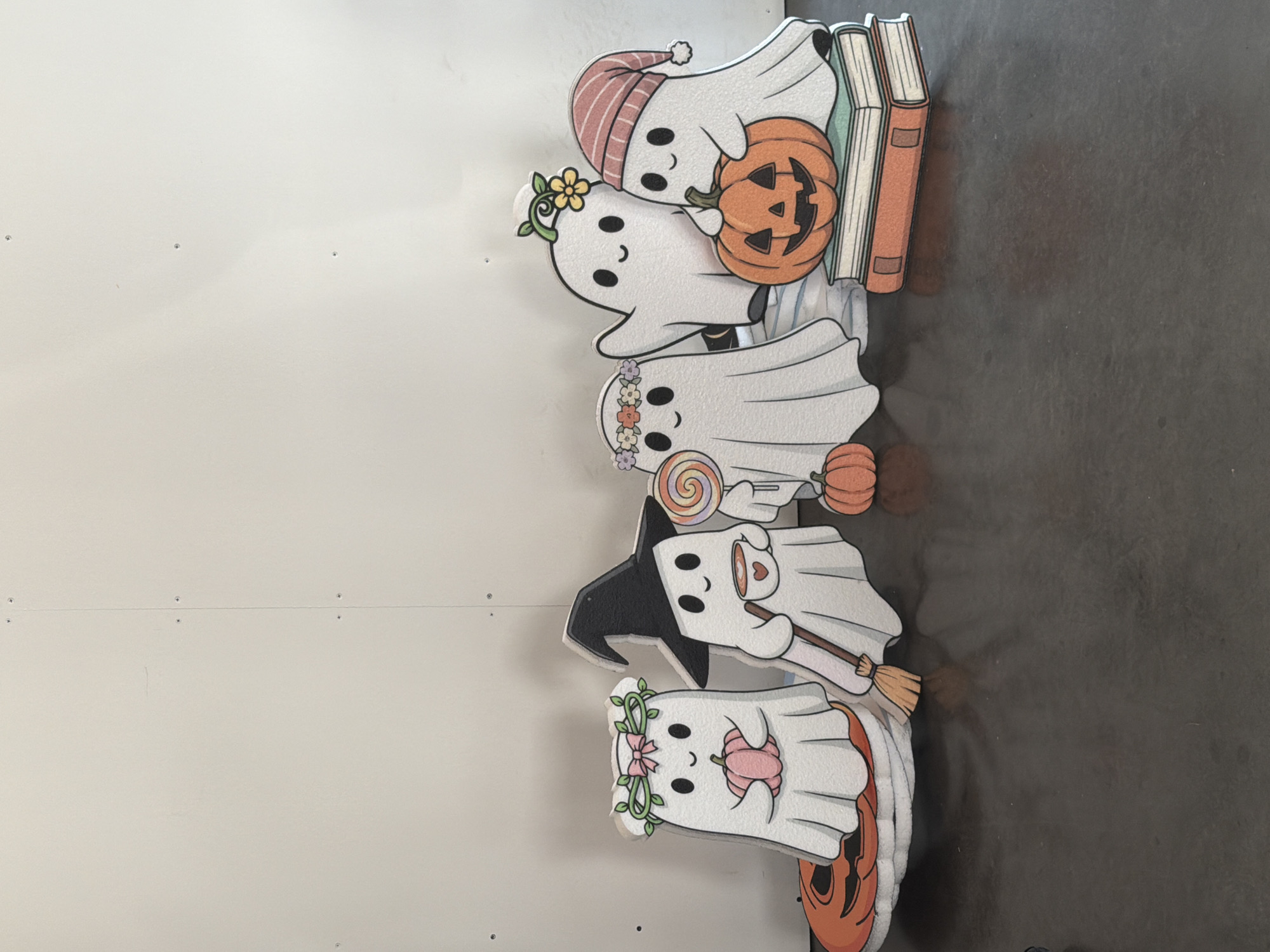 A group of five cartoon ghosts, designed as Halloween decorations, are arranged on a dark, reflective surface in front of a plain, light-colored wall. The ghosts are depicted in a flat, cutout style with simple facial features.
From left to right:
1. A ghost wearing a green wreath with a pink bow and holding a pink pumpkin. It stands on a circular orange and black object resembling a carved pumpkin.
2. A ghost wearing a black witch's hat and holding a mug with a heart design. A broomstick is positioned next to it.
3. A ghost wearing a flower crown, holding a swirled lollipop.
4. A ghost with its arm raised in a waving gesture.
5. A ghost wearing a pink striped hat and resting its hands on a carved pumpkin that sits on top of two books. A small, uncarved orange pumpkin is also visible to the left of this ghost.
The wall behind the ghosts has visible seams and small, dark circular indentations, suggesting it might be a drywall surface with screw heads or fasteners. The lighting appears to be from above and slightly in front, casting subtle shadows on the floor. The overall scene is staged for a festive, possibly seasonal, display.