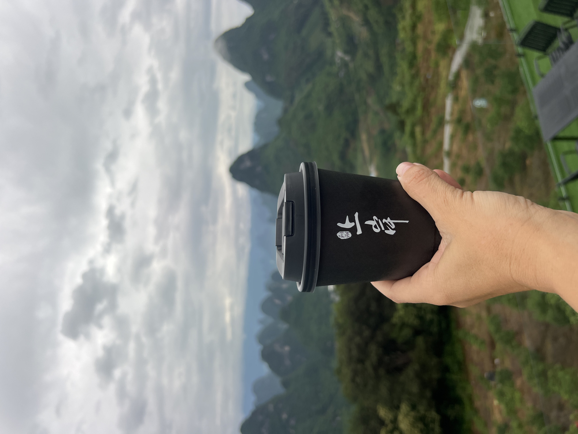 A hand holds a black travel mug with white Chinese characters. The background shows a mountainous landscape under a cloudy sky. The location is Guilin, China, based on the karst mountains.