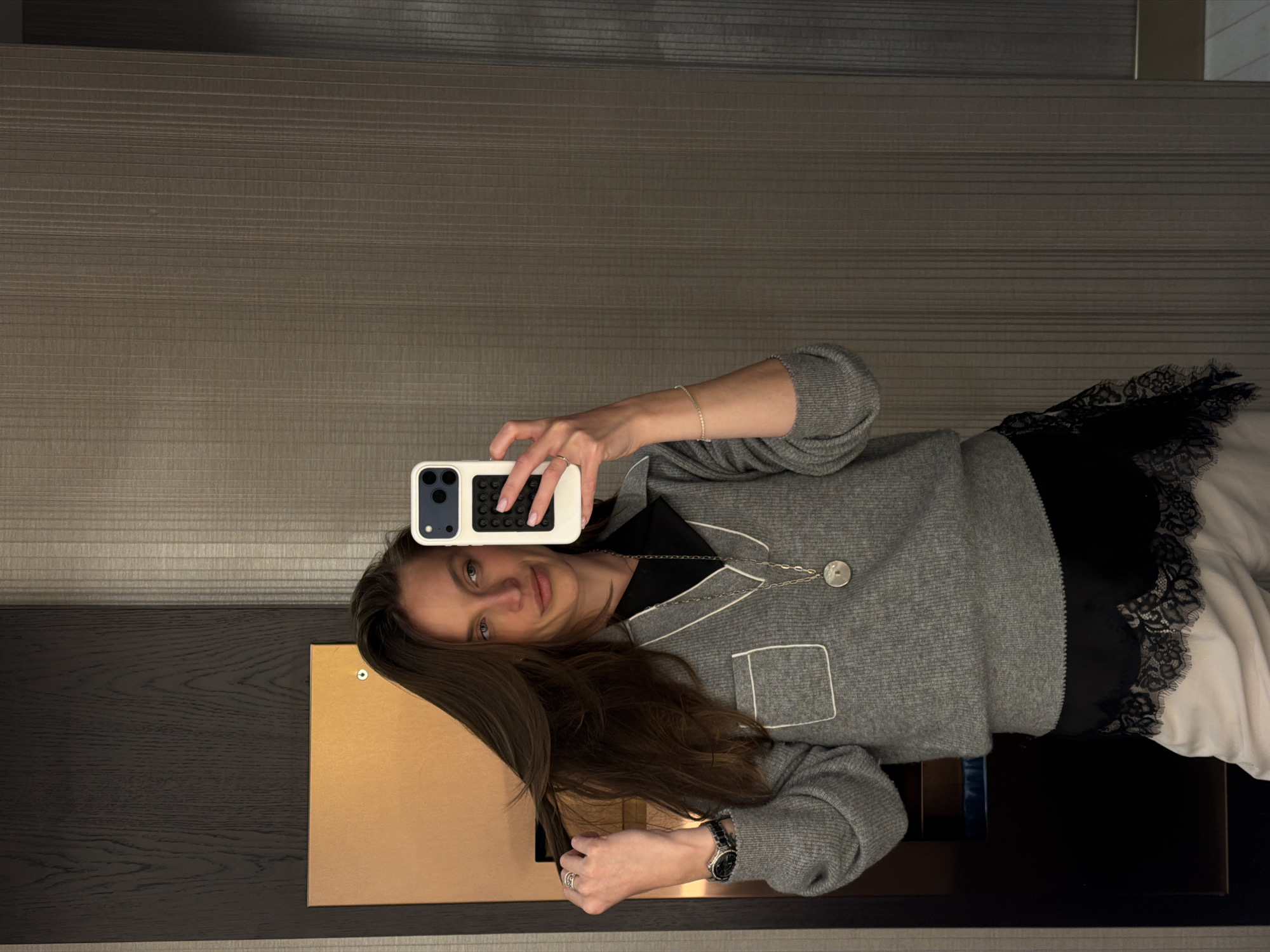 A woman is holding a smartphone in her right hand, taking a selfie. She is wearing a gray sweater with white trim and a black and white lace-trimmed garment. The background consists of a textured wall above a dark-colored cabinet with a light-colored panel on it. Based on context clues, the location might be Budva, Montenegro.