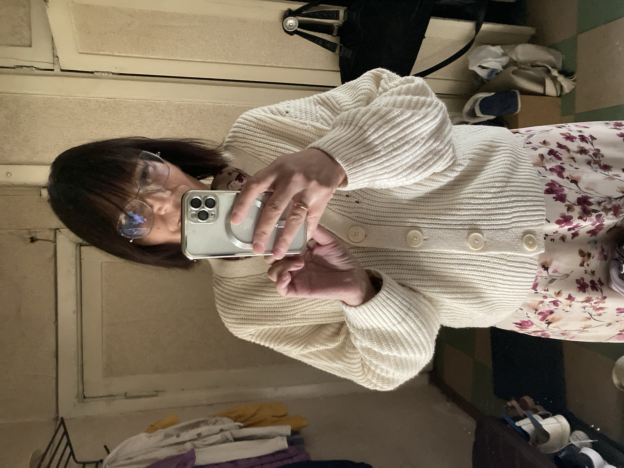A medium shot captures a woman taking a mirror selfie in what appears to be a cluttered entryway or hallway. She is wearing a cream-colored cable-knit cardigan with three buttons and a floral print skirt with pink and purple flowers. She has short dark hair and is wearing glasses. In her right hand, she holds a smartphone, angled to capture her reflection. Behind her, a rack of clothes is visible, along with a closed door and a bag hanging on a hook. The floor is tiled with a geometric pattern. Shoes are visible on the floor and a small shelf. The setting suggests a residential interior, possibly in Novokuznetsk, Russia, given the context provided.