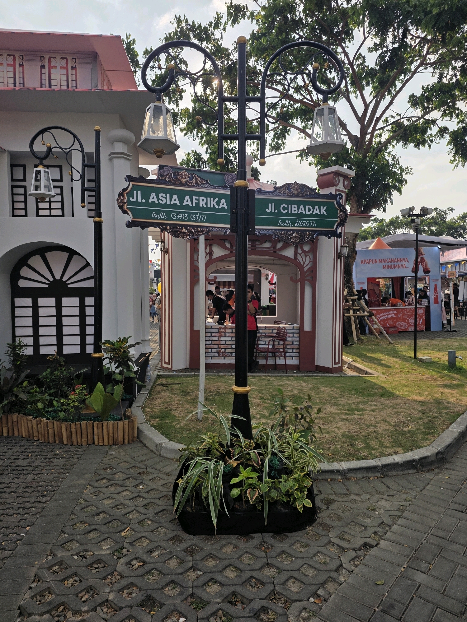 The image shows a street signpost in the city of Bandung, Indonesia, indicating "Jl. Asia Afrika" and "Jl. Cibadak". The signpost is ornate, with a black pole and decorative gold accents, and features two hexagonal lanterns. It is surrounded by greenery, with plants in a planter at the base of the pole. In the background, there is a white building with arched windows and a red roof, and beyond that, an outdoor market or event space with tents and booths. Several people are visible in the background, some appearing to be customers or vendors at the stalls. The ground is paved with interlocking concrete blocks. The sky is visible at the top of the image, with some clouds and sunlight filtering through tree branches.
