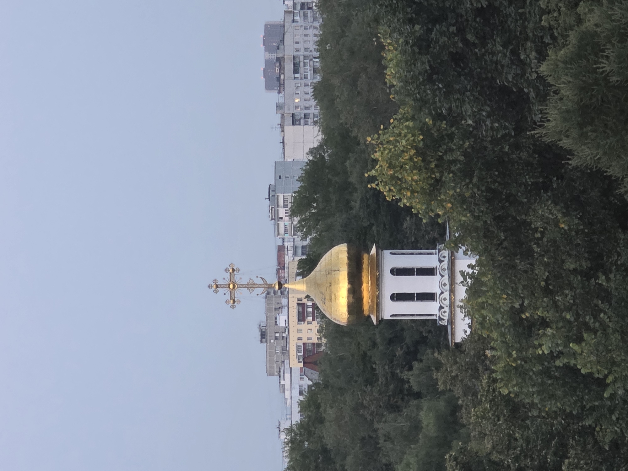 The image depicts an Orthodox church dome with a golden cross, rising above a dense canopy of trees. In the background, several multi-story apartment buildings are visible, characteristic of urban Russian architecture. The sky is overcast and muted in color, suggesting it might be early morning or late evening. The specific location within Moscow is not identifiable from the visual cues alone. There are no people or discernible actions in the scene. The overall atmosphere is serene and contemplative.