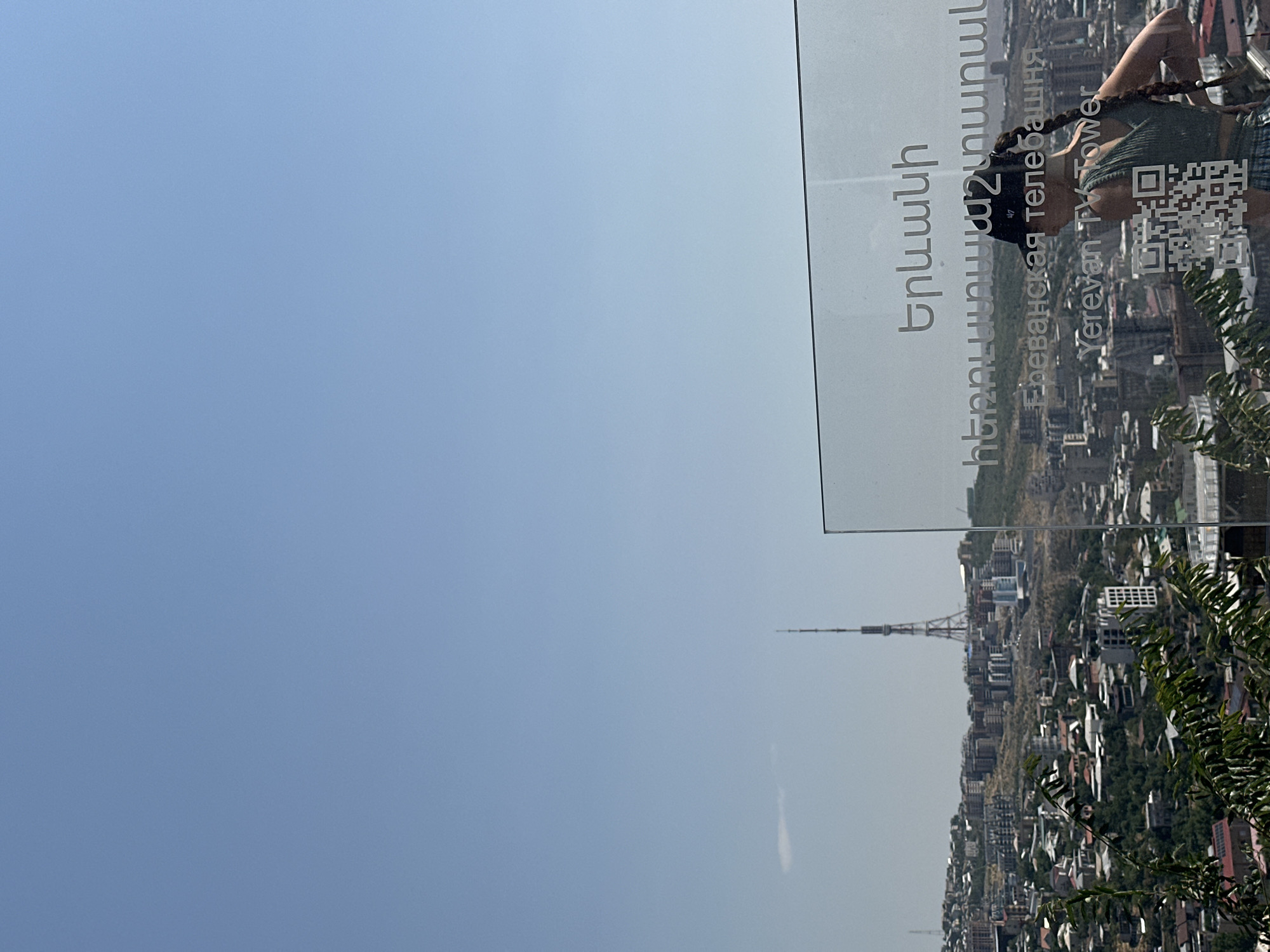 A medium shot captures a woman with long braided hair, wearing a black baseball cap and a green tank top, standing on a raised viewpoint overlooking the city of Yerevan, Armenia. She is facing away from the camera, with her right hand on her hip and her left arm bent, as if adjusting her cap. In the foreground, a transparent glass barrier with text inscribed on it reflects the cityscape. The text, visible in Armenian, Russian, and English, identifies the location as "Yerevan TV Tower." A QR code is also etched onto the glass. The background reveals a sprawling urban landscape with numerous buildings of varying architectural styles spread across hilly terrain. The Yerevan TV Tower, a prominent latticed structure, stands tall in the distance. The sky above is clear and blue.