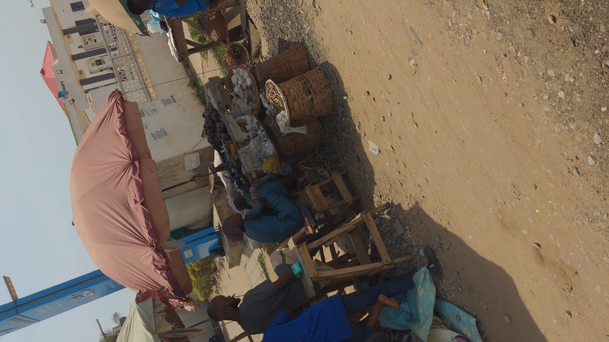 The image captures a vibrant street market scene in Ado, Nigeria, observed from a slightly elevated and tilted perspective. The setting features a bustling roadside area with vendors and a dirt and gravel road in the foreground and right side.
In the center, a makeshift market stall is set up, shaded by a large, reddish-brown fabric umbrella. Various goods are displayed on and around low wooden structures and woven baskets. These goods include baskets filled with what appear to be eggs or small packaged items, another large basket containing nuts or grains, and a pile of dark, oval-shaped fruits, possibly avocados or mangoes. Scattered plastic bags and other small items are also visible.
Several individuals are present, engaged in daily commerce. A person in a blue patterned outfit is seated on a low wooden stool near the market goods. Another man with a shaved head and beard, wearing a grey t-shirt and dark trousers, is seated on a similar low wooden structure in the foreground, seemingly occupied with something in his lap. Parts of other individuals in blue clothing are visible near the stalls.
In the background, a white building with a red-tiled roof is clearly identifiable as a petrol station, with fuel pumps visible in front. The letters "NPC" are discernible on the building's facade. Small, illegible text appears on the white wall below the petrol station.
The scene takes place during the daytime, indicated by the bright light and strong shadows cast by the umbrella and people, suggesting either morning or late afternoon. The sky is clear. The overall atmosphere depicts a typical, functional street market in an urban or semi-urban African context.