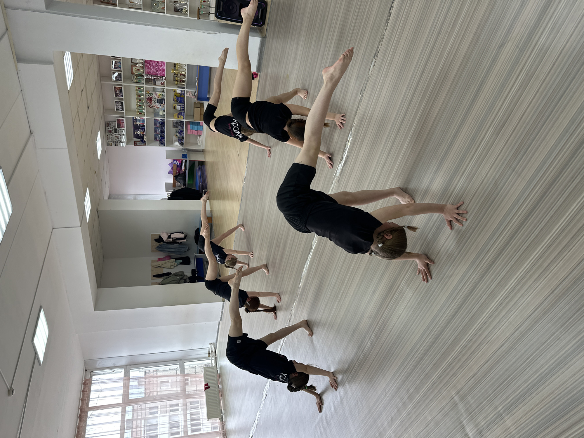 In an indoor gymnasium in Votkinsk, Russia, a line of five young female gymnasts practice a routine. The gymnasts, all wearing black shirts and shorts, are spaced apart on a light-colored, striped floor. They are performing a handstand-like pose with one leg extended upwards and the other extended forward. The gymnasium has white walls, fluorescent lighting, and a window with a radiator below it. Along the back wall, there are shelves displaying various items including trophies, photographs, and other objects.